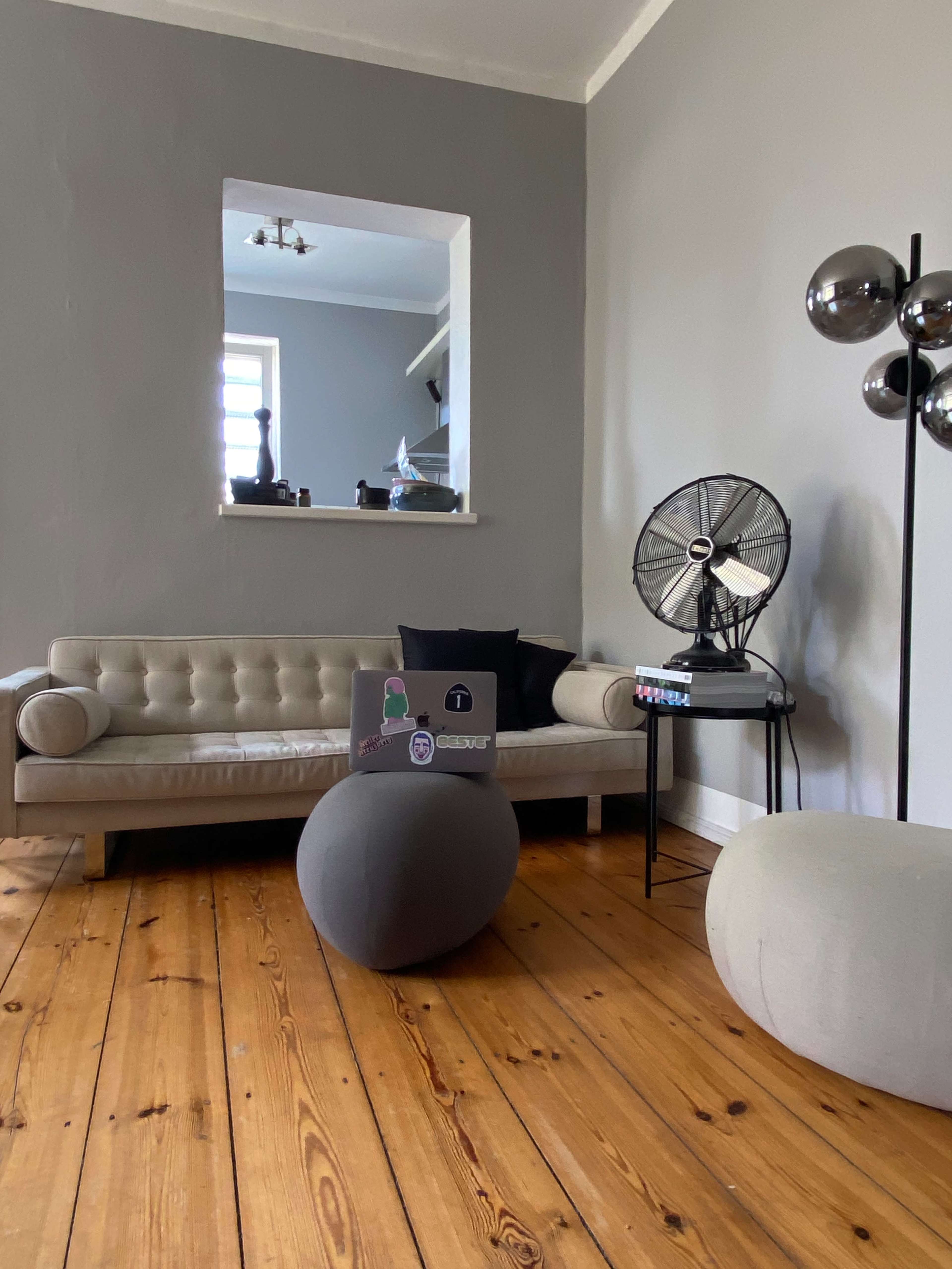 A minimalist living room features a beige tufted sofa, a gray round table, and a standing fan on a black side table, all situated on a wooden floor.