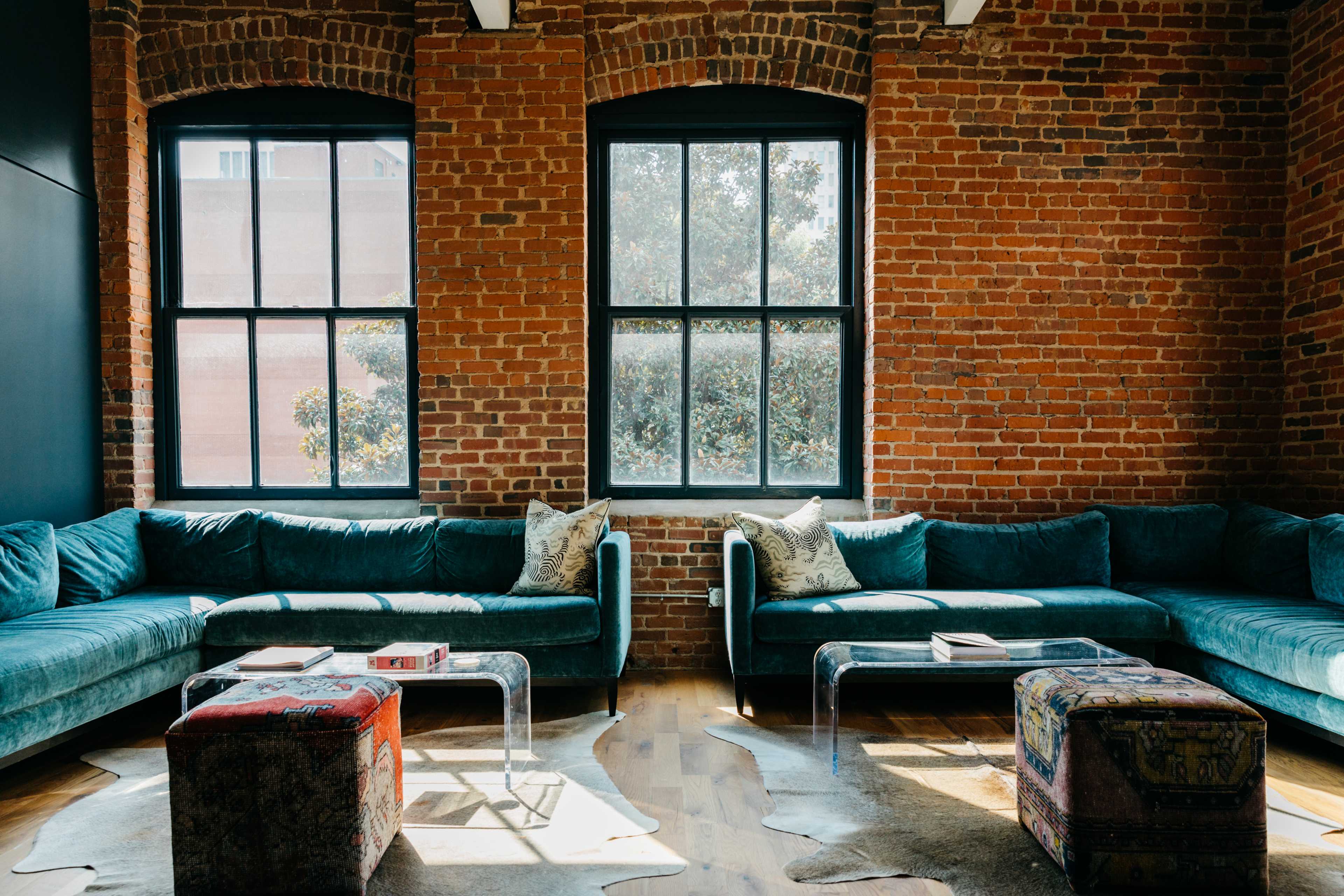 A spacious room with two teal sofas facing each other, large windows with brick walls, and a glass coffee table on a wooden floor.