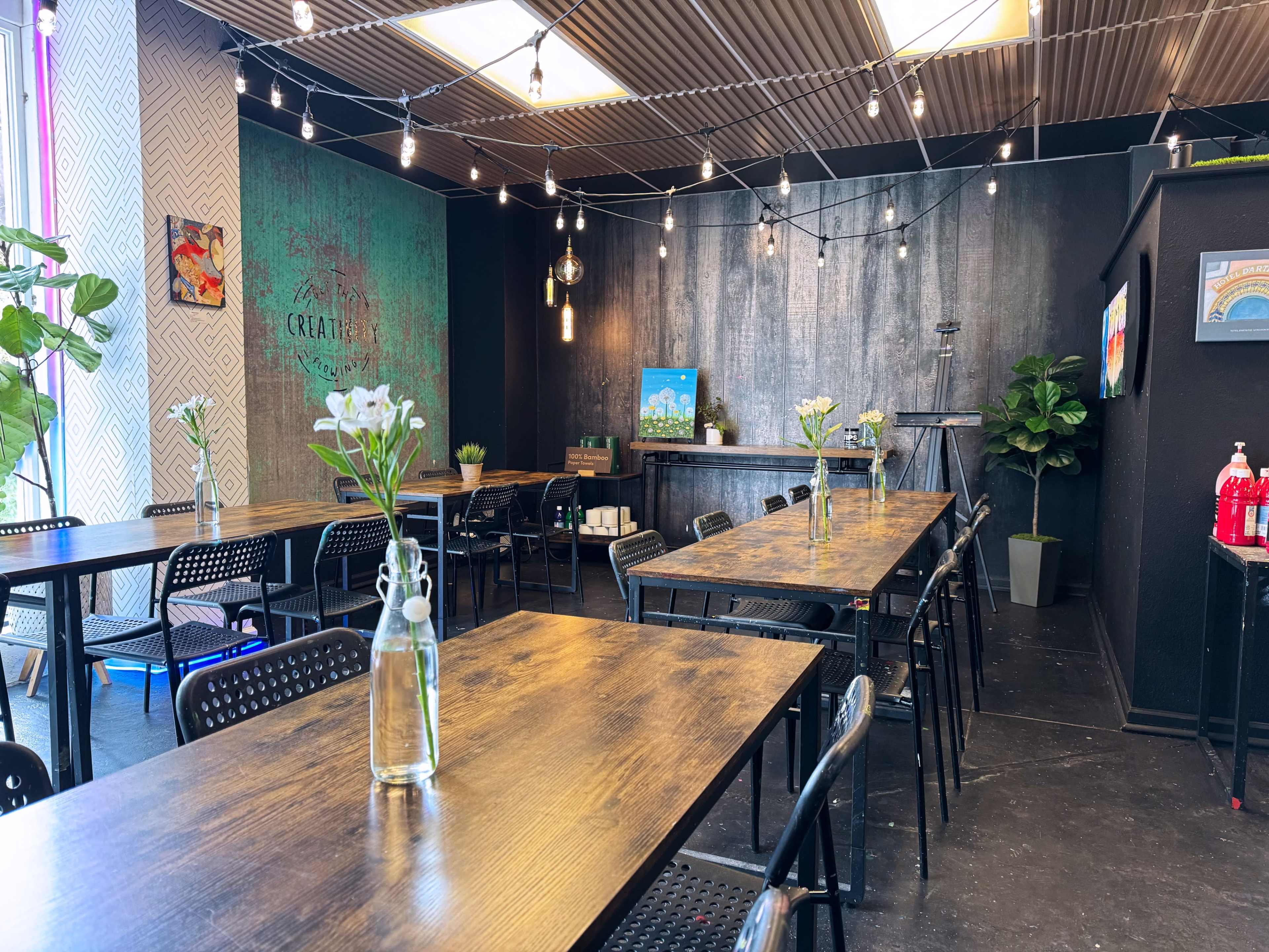 The image shows a decorated interior of a café with wooden tables, black chairs, and string lights overhead.