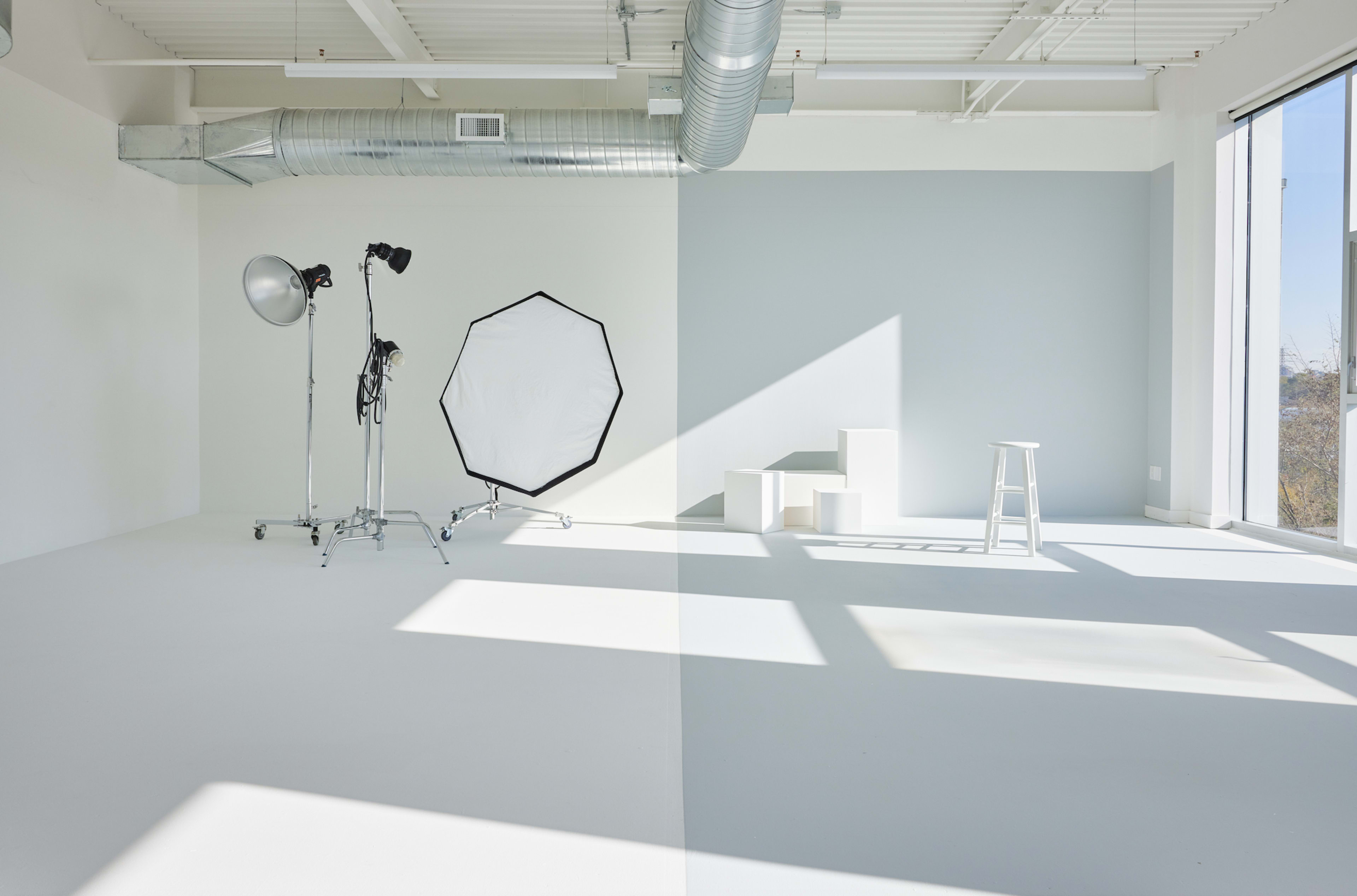 A bright photography studio with a large softbox, a stand for a light, and a small white stool, all positioned on a light-colored floor.