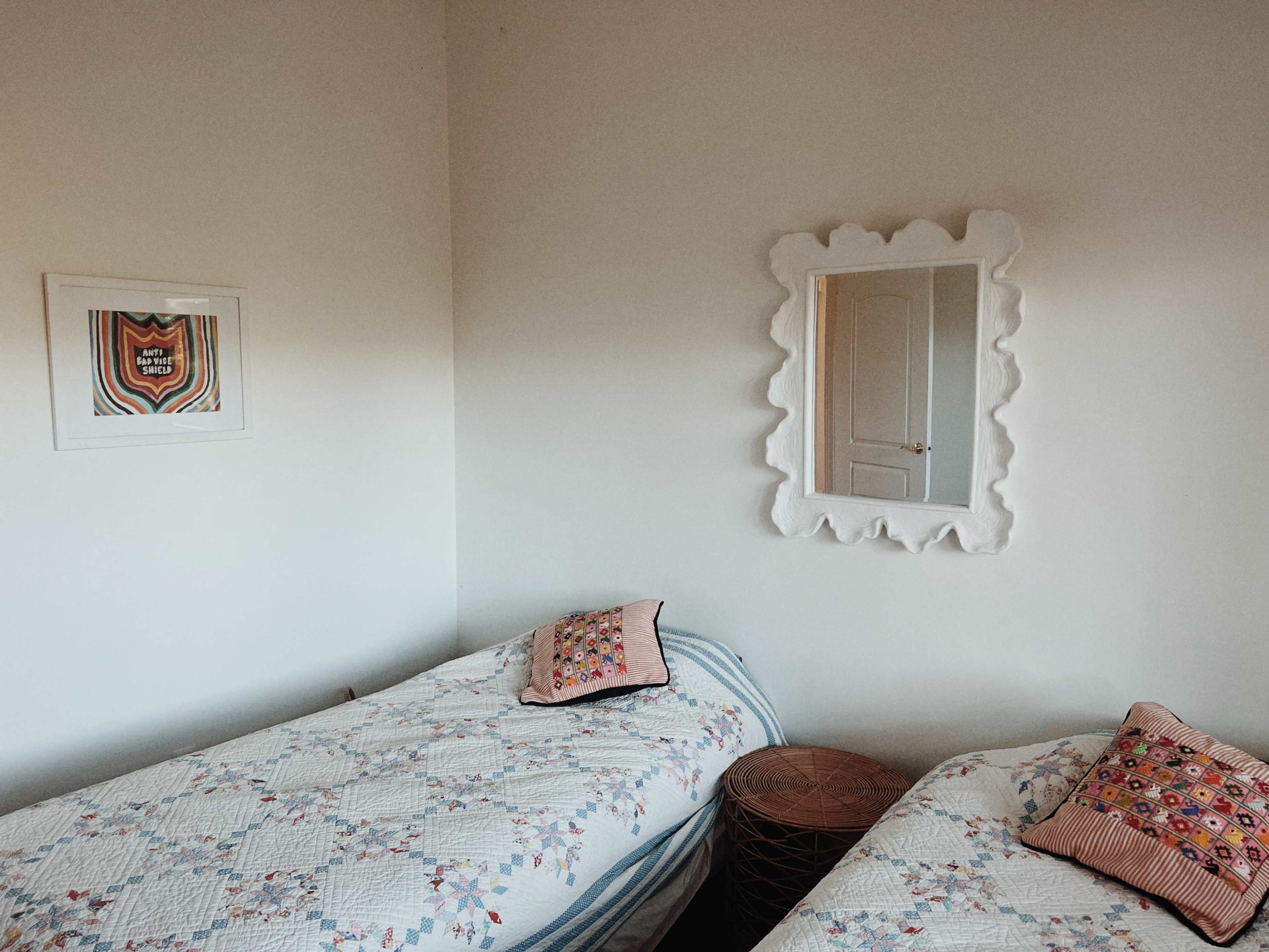 The image shows a well-lit bedroom with two twin beds dressed in patterned quilts, a textured mirror on the wall, and a decorative print framed nearby.