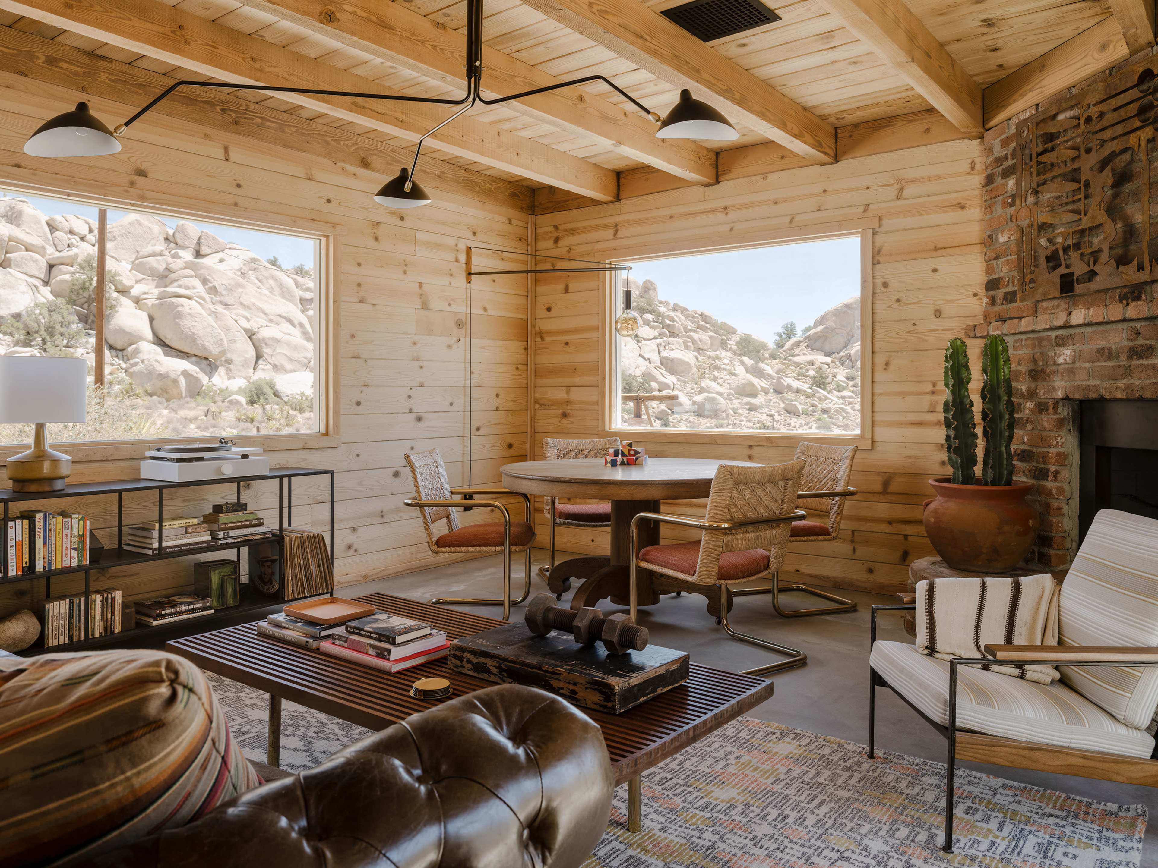 The image shows a wooden interior living space with a round table, bookshelf, and a seating area featuring a leather couch and armchairs, all surrounded by large windows that offer a view of rocky terrain outside.