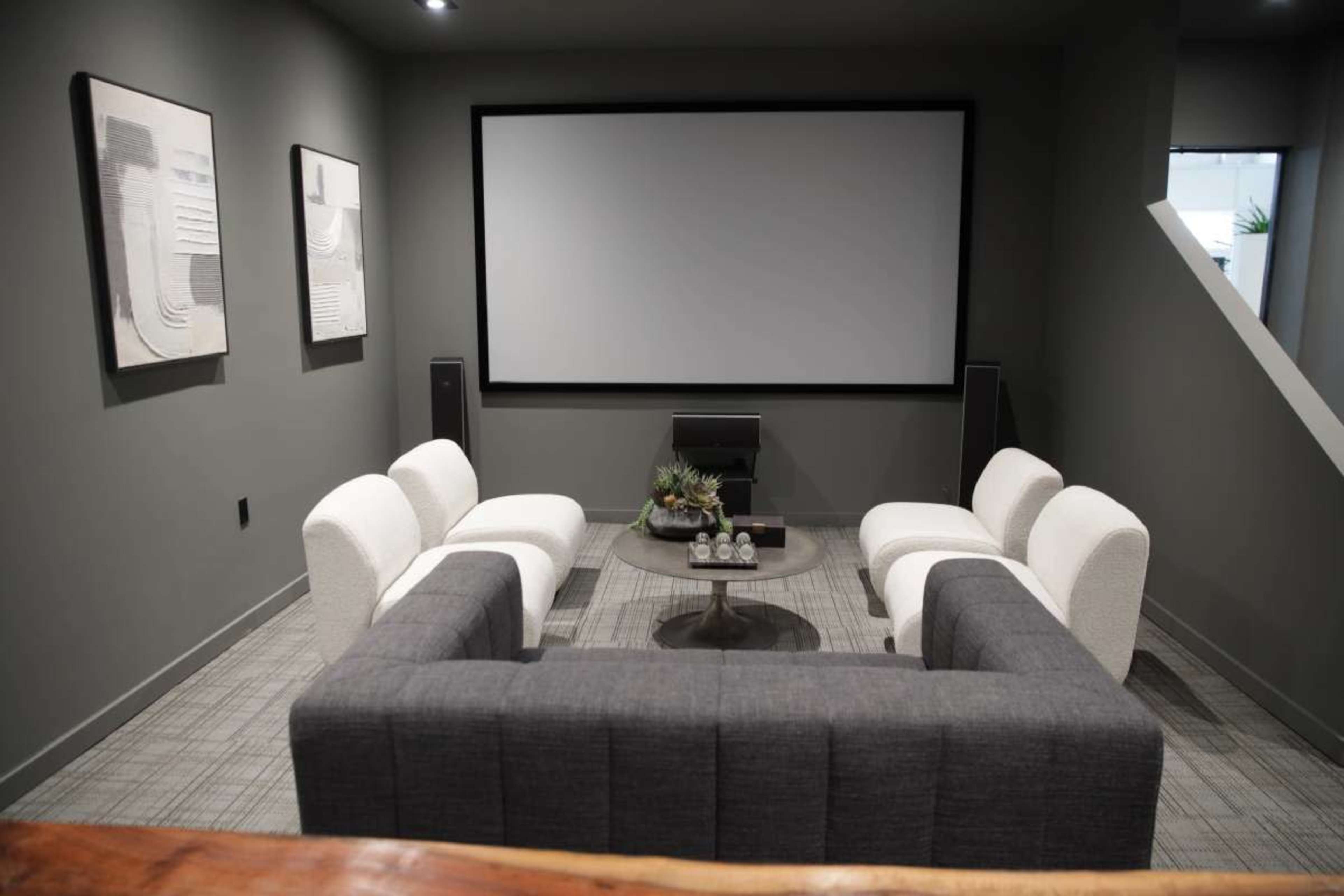 The image shows a modern living room with a large screen against the wall, surrounded by various seating arrangements and a coffee table in the center.