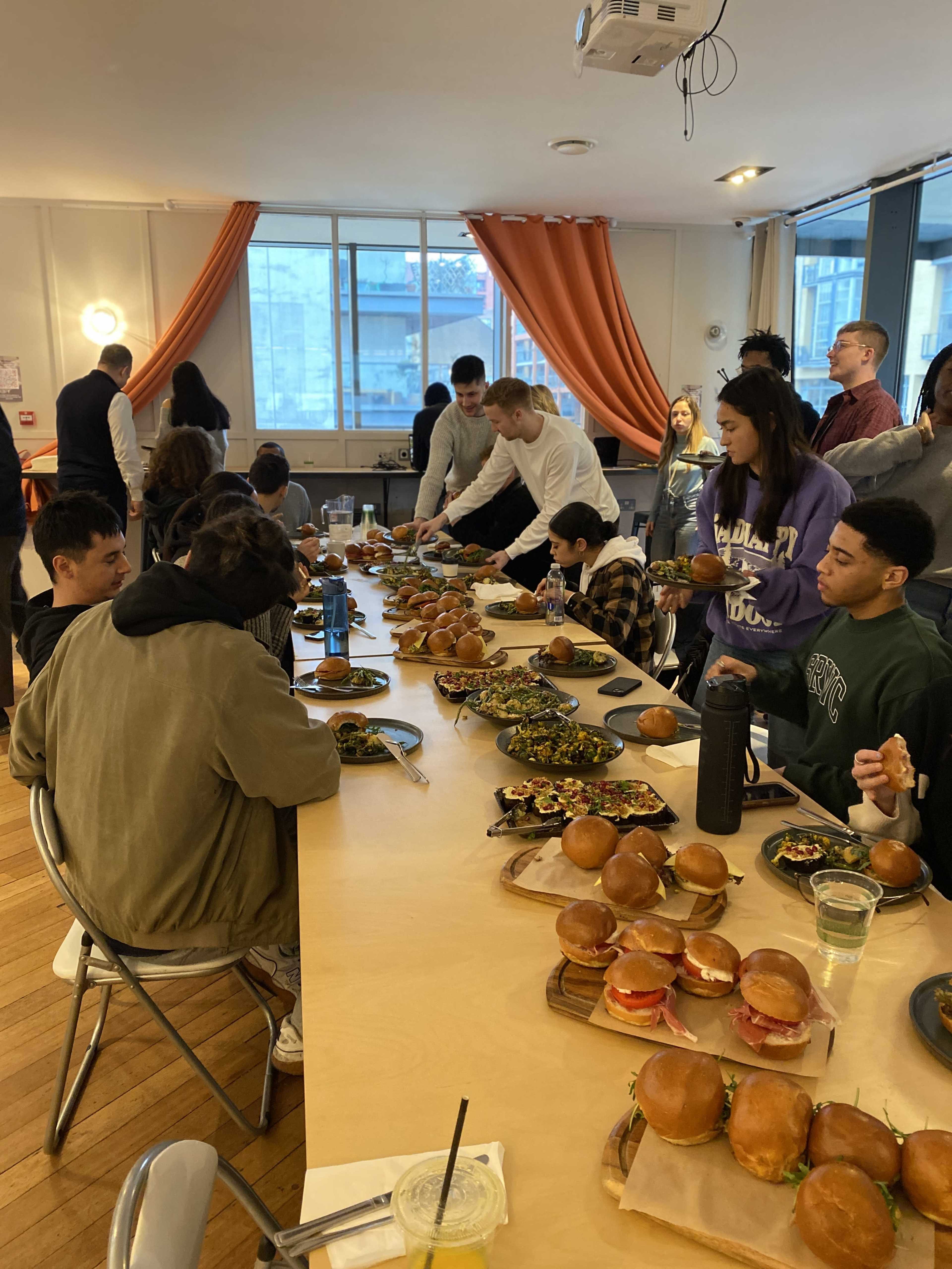 A group of people is gathered around a long table filled with burgers and salads in a well-lit room.