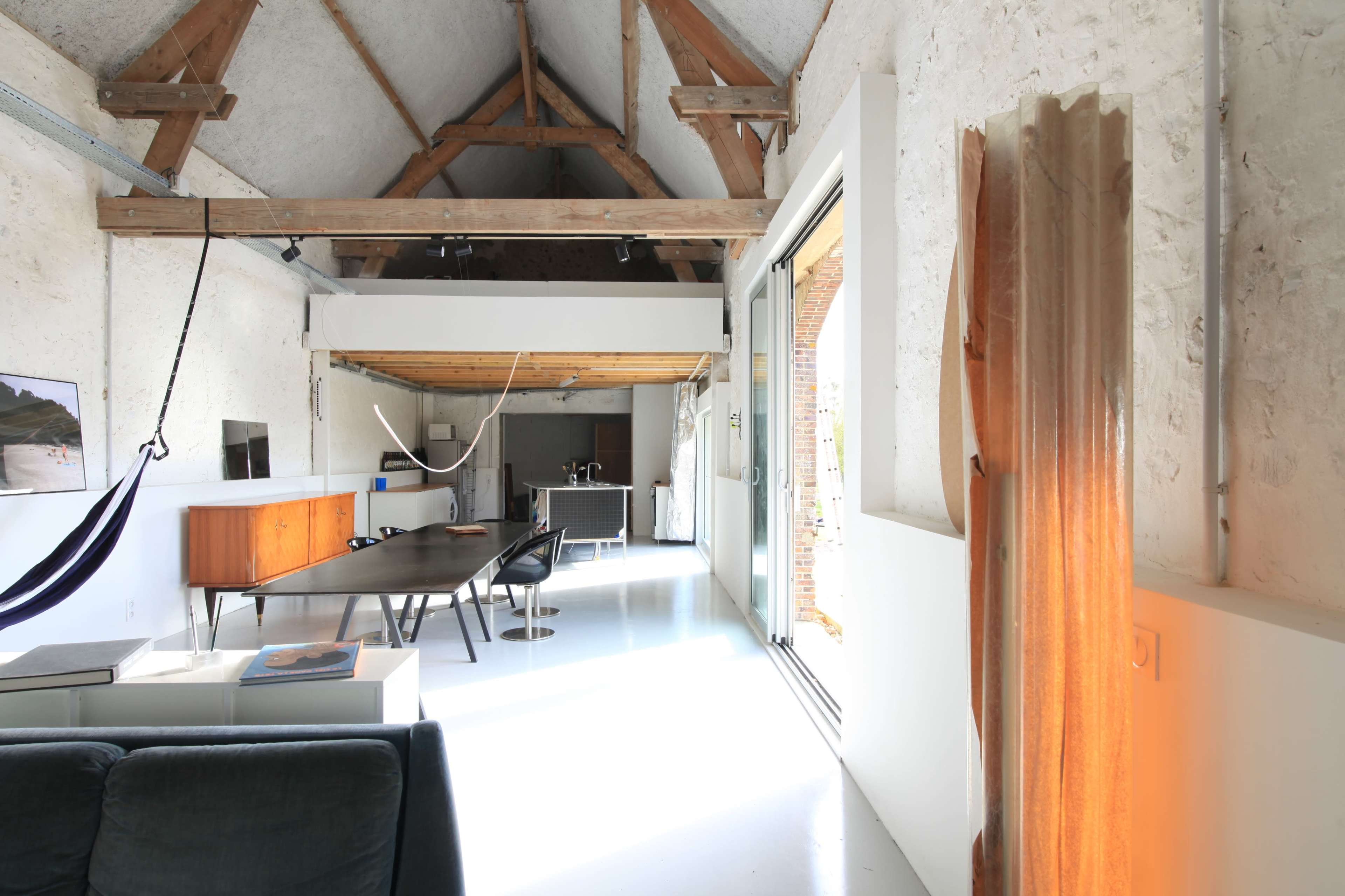 A bright, spacious interior of a modern living area with a wooden beam ceiling, a dining table, a hammock, and large windows.