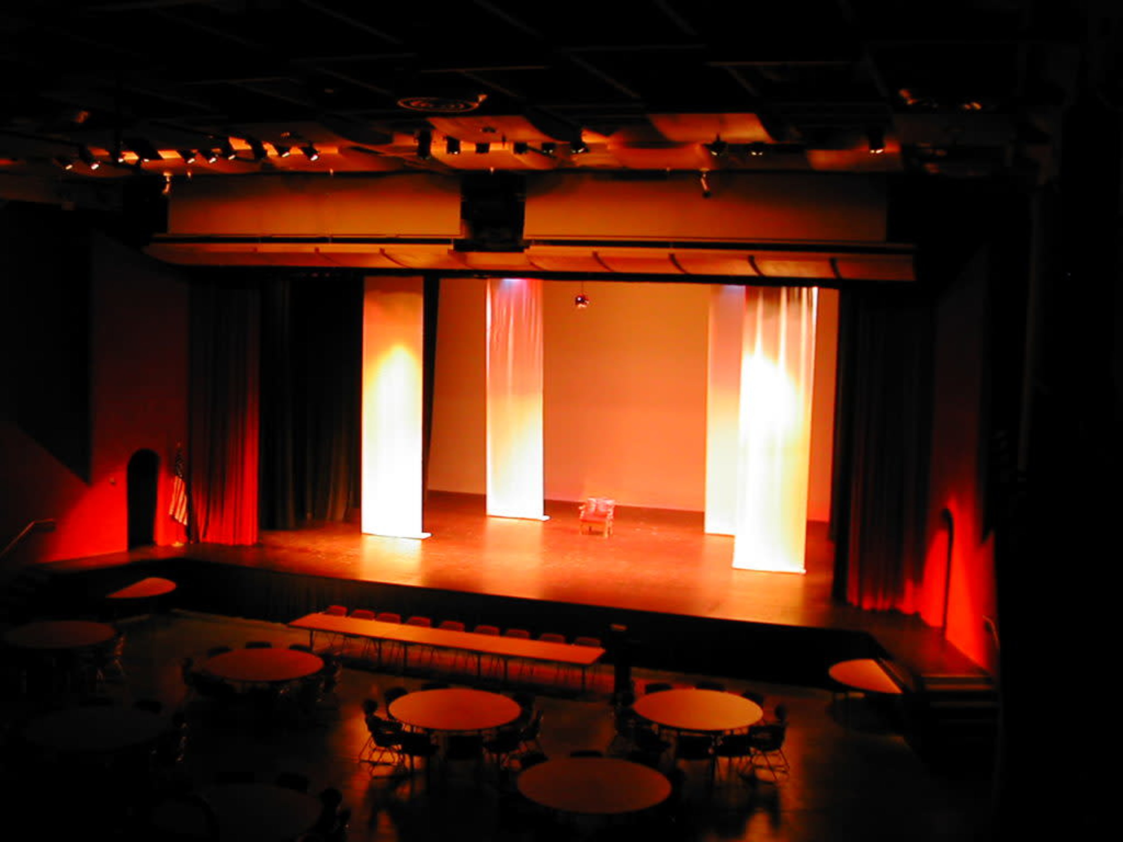 The image shows an empty stage with two tall white curtains and a row of tables and chairs set up in the audience area.
