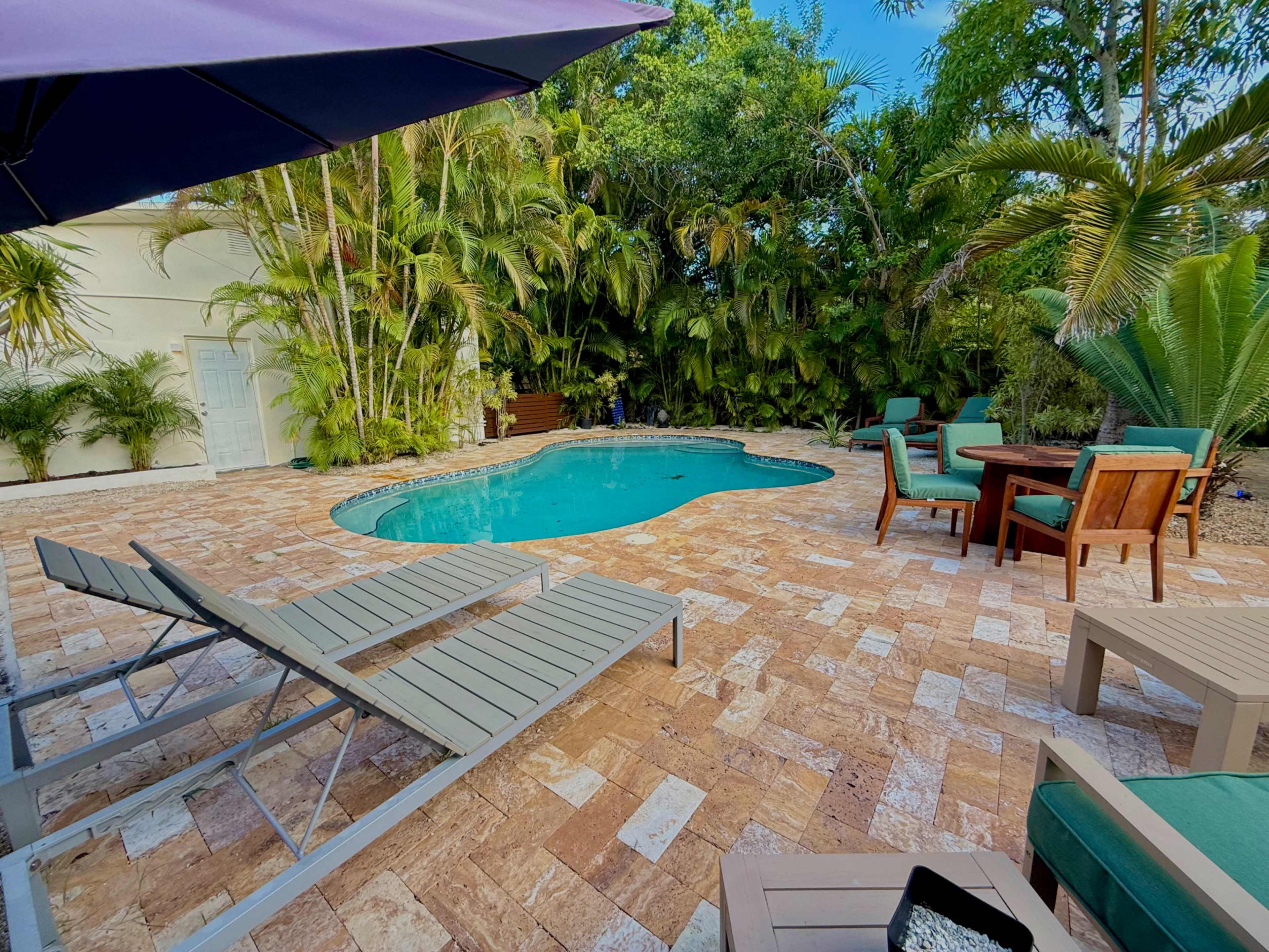 A swimming pool surrounded by deck chairs and outdoor furniture is set in a tropical landscape with lush greenery.