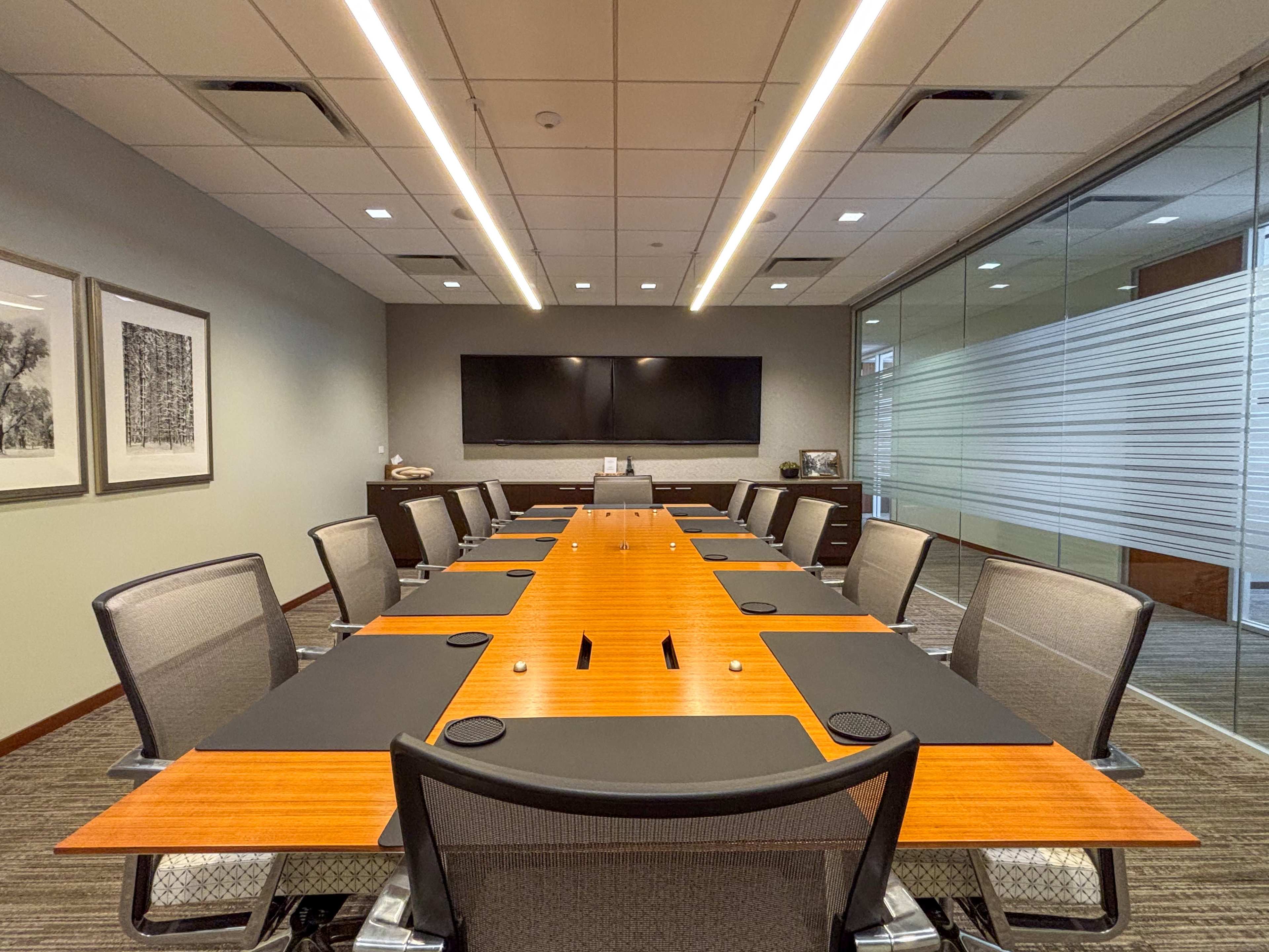 A modern conference room features a long wooden table surrounded by ergonomic chairs, with a large screen on the wall and glass partitions.