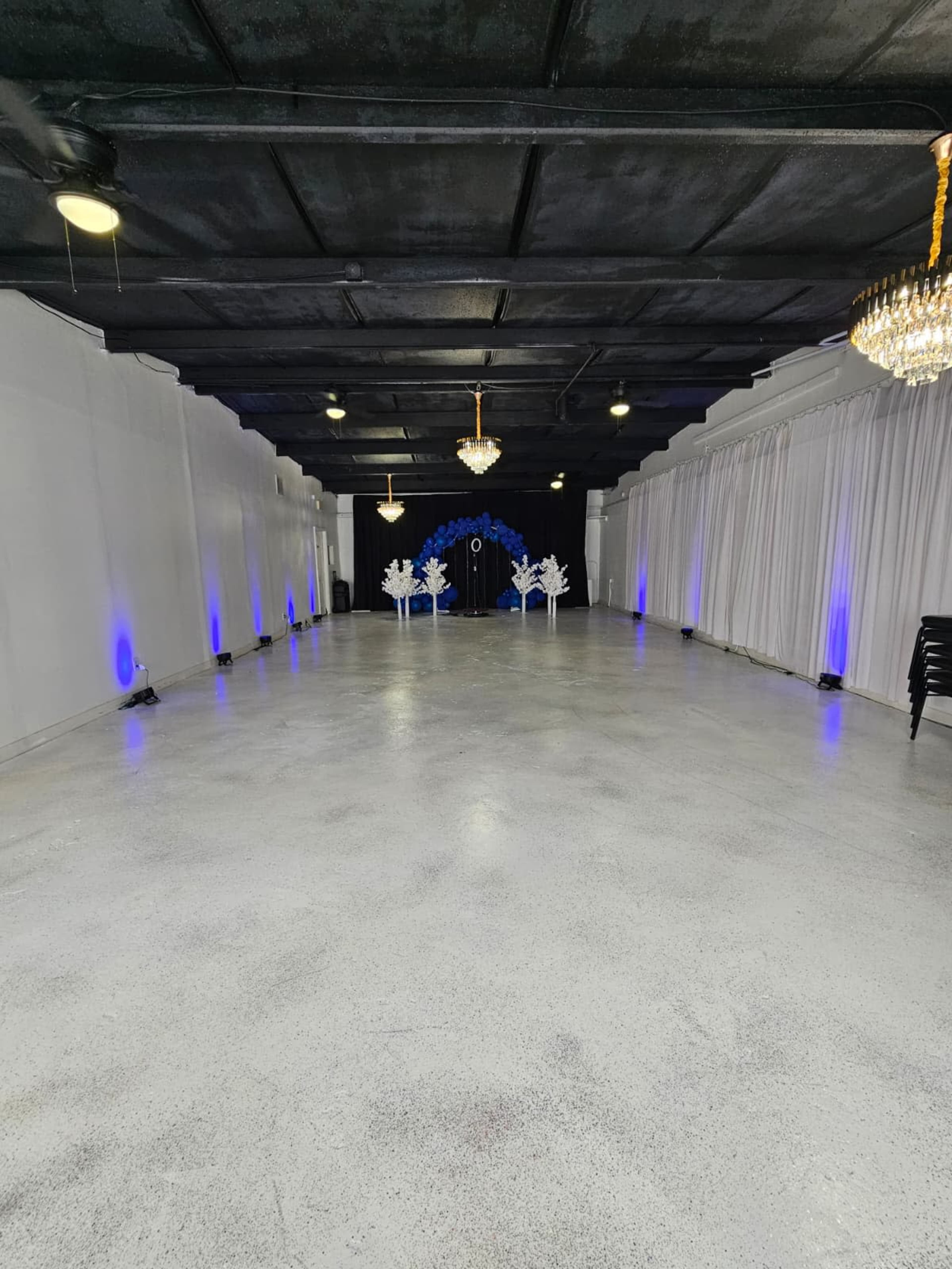 The image shows a spacious, empty event hall with a polished concrete floor, white drapery along the walls, and blue lighting illuminating the area.