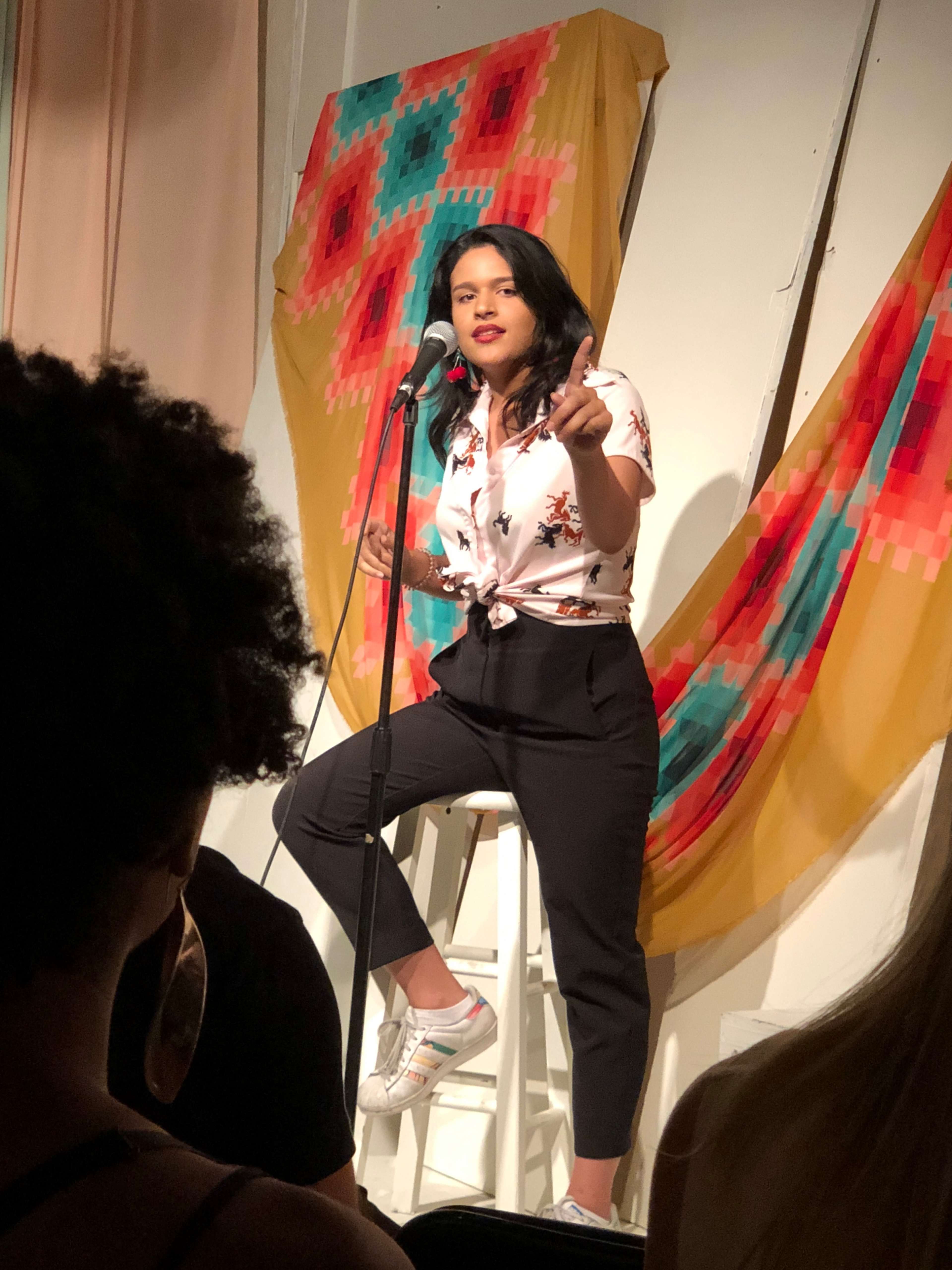 A performer stands on a stool, speaking into a microphone in front of a colorful backdrop during a comedy show.