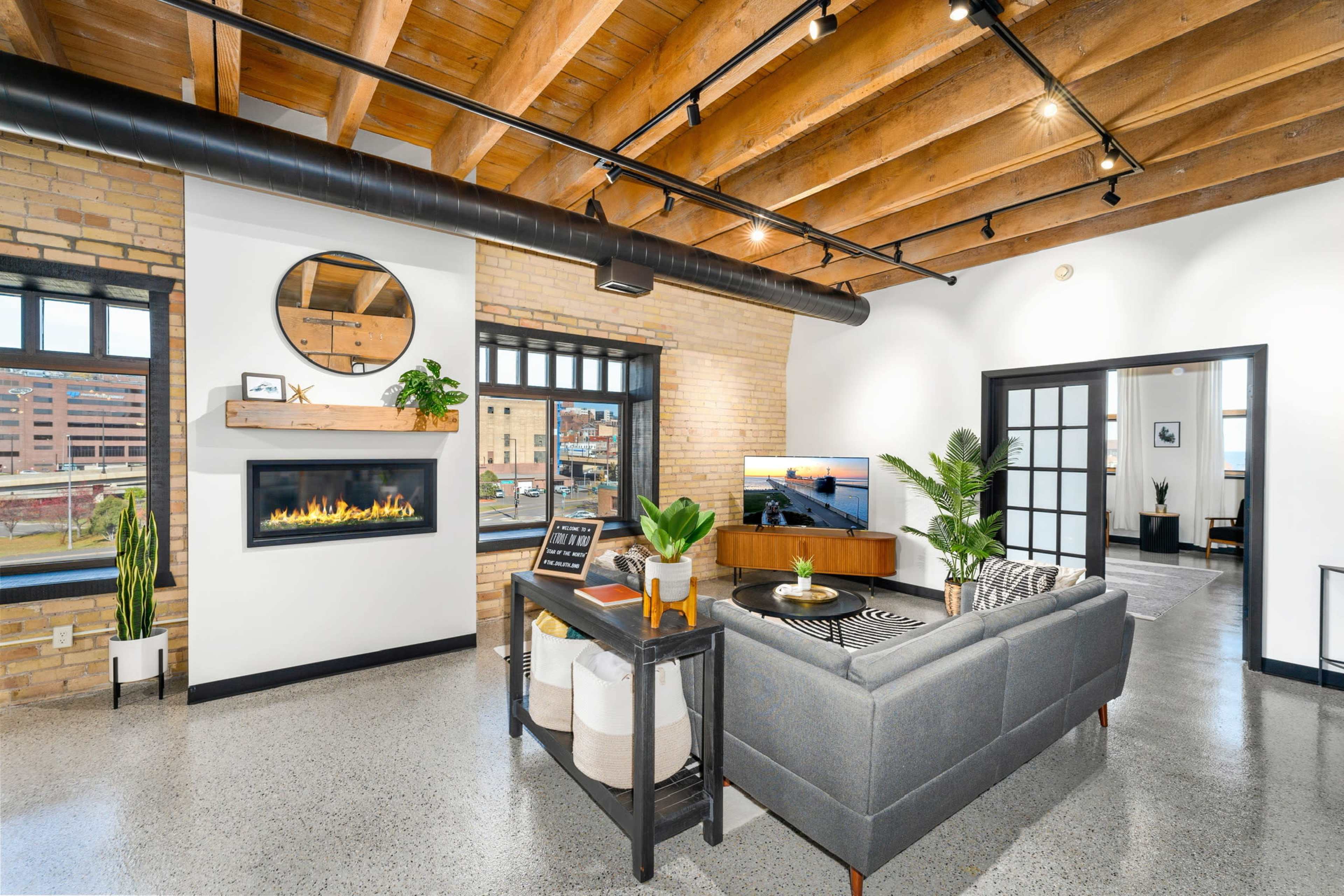 A modern living room with a gray sofa, a mounted television, a fireplace, and large windows, featuring exposed wooden beams and polished concrete flooring.