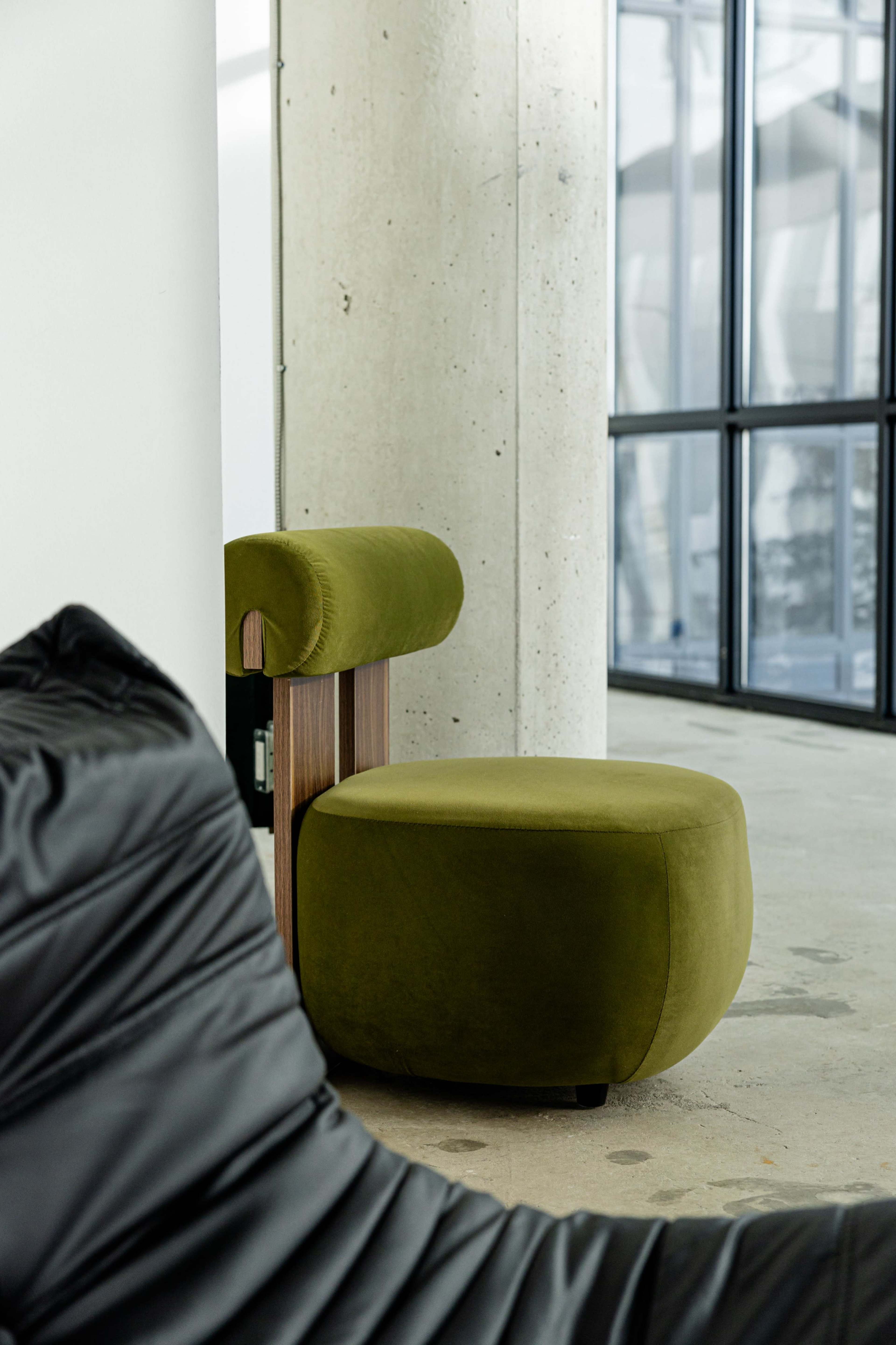The image features a green upholstered chair with a wooden base positioned next to a black bean bag in a modern interior space with large windows.
