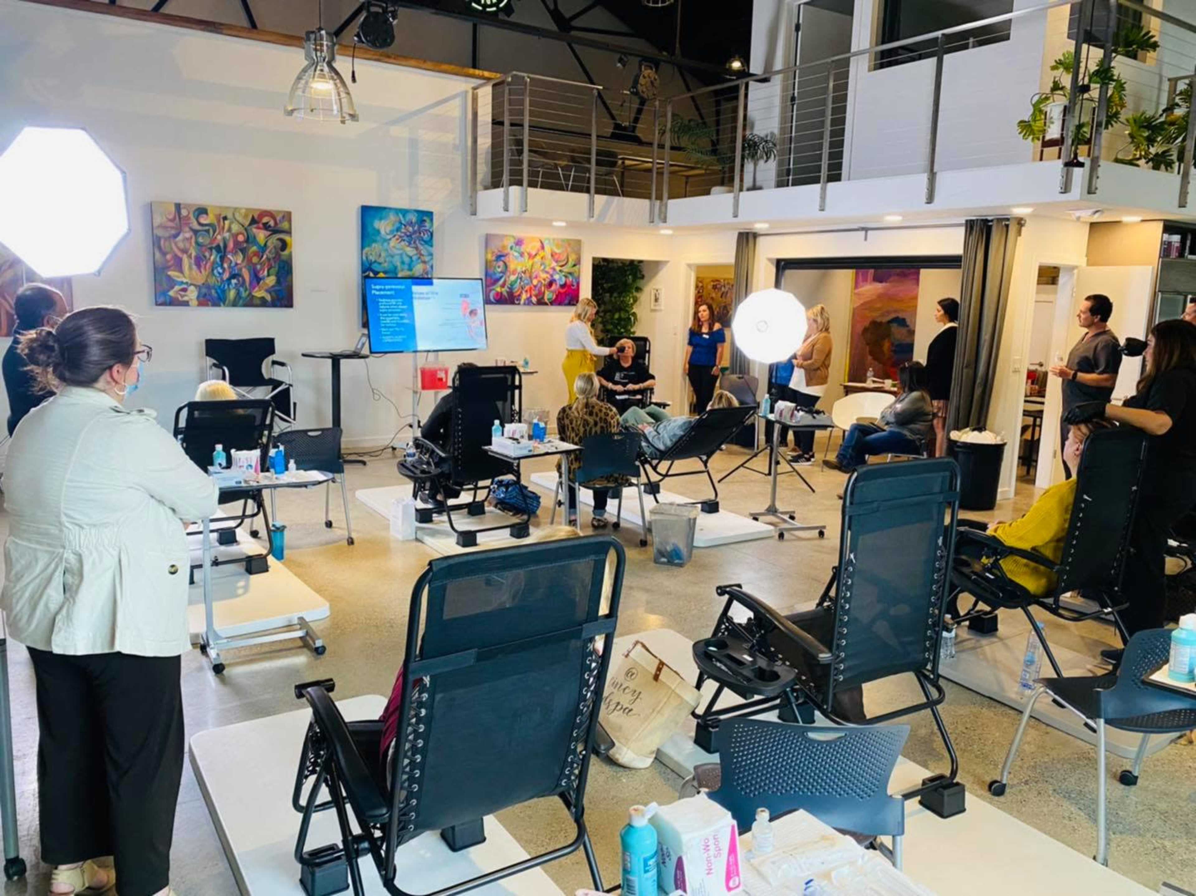 A group of people gathers in a modern, well-lit studio with salon chairs and art on the walls for a demonstration or event.