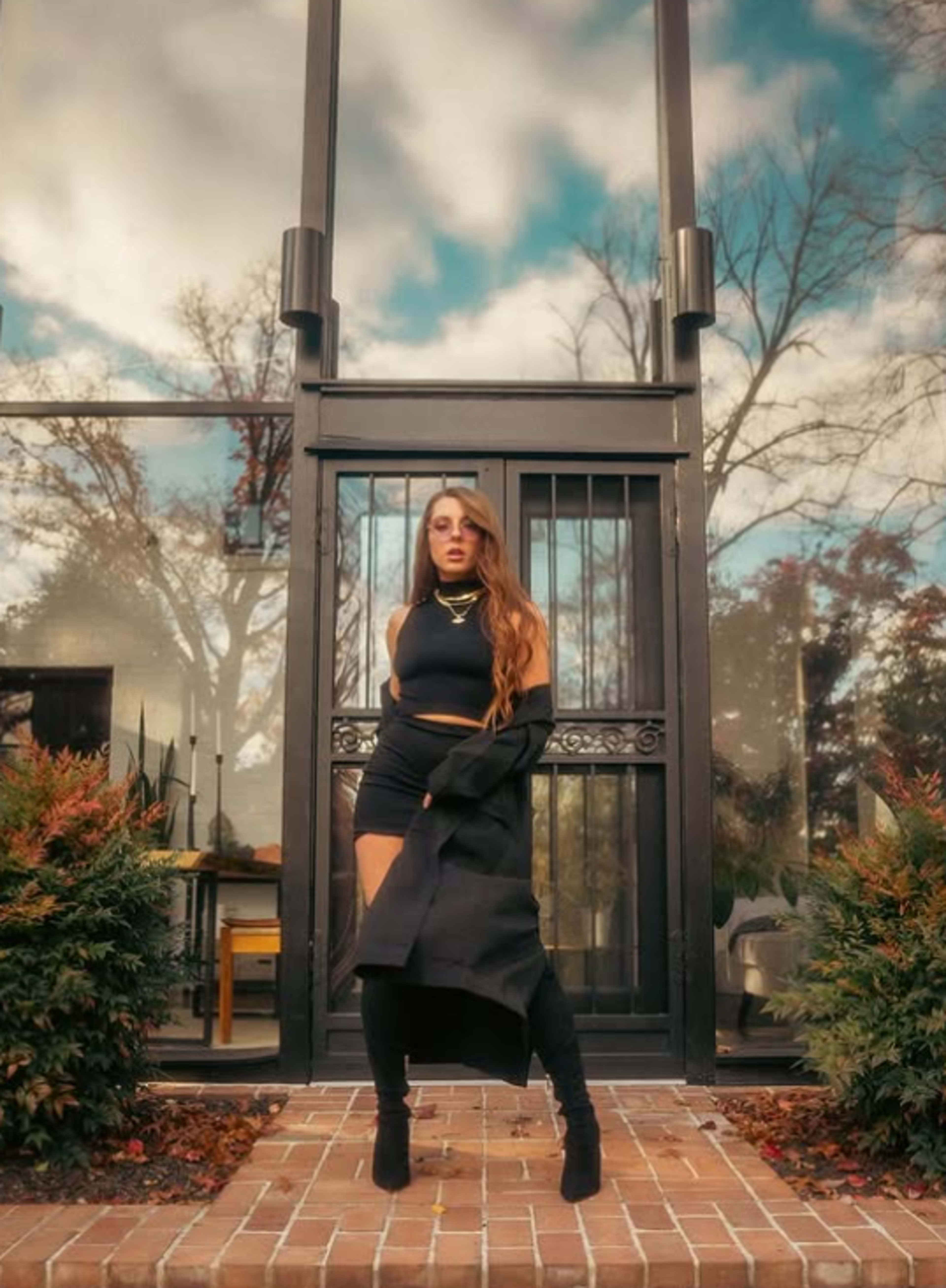A woman with long hair stands confidently on a brick pathway in front of a glass door, wearing a black outfit and knee-high boots.