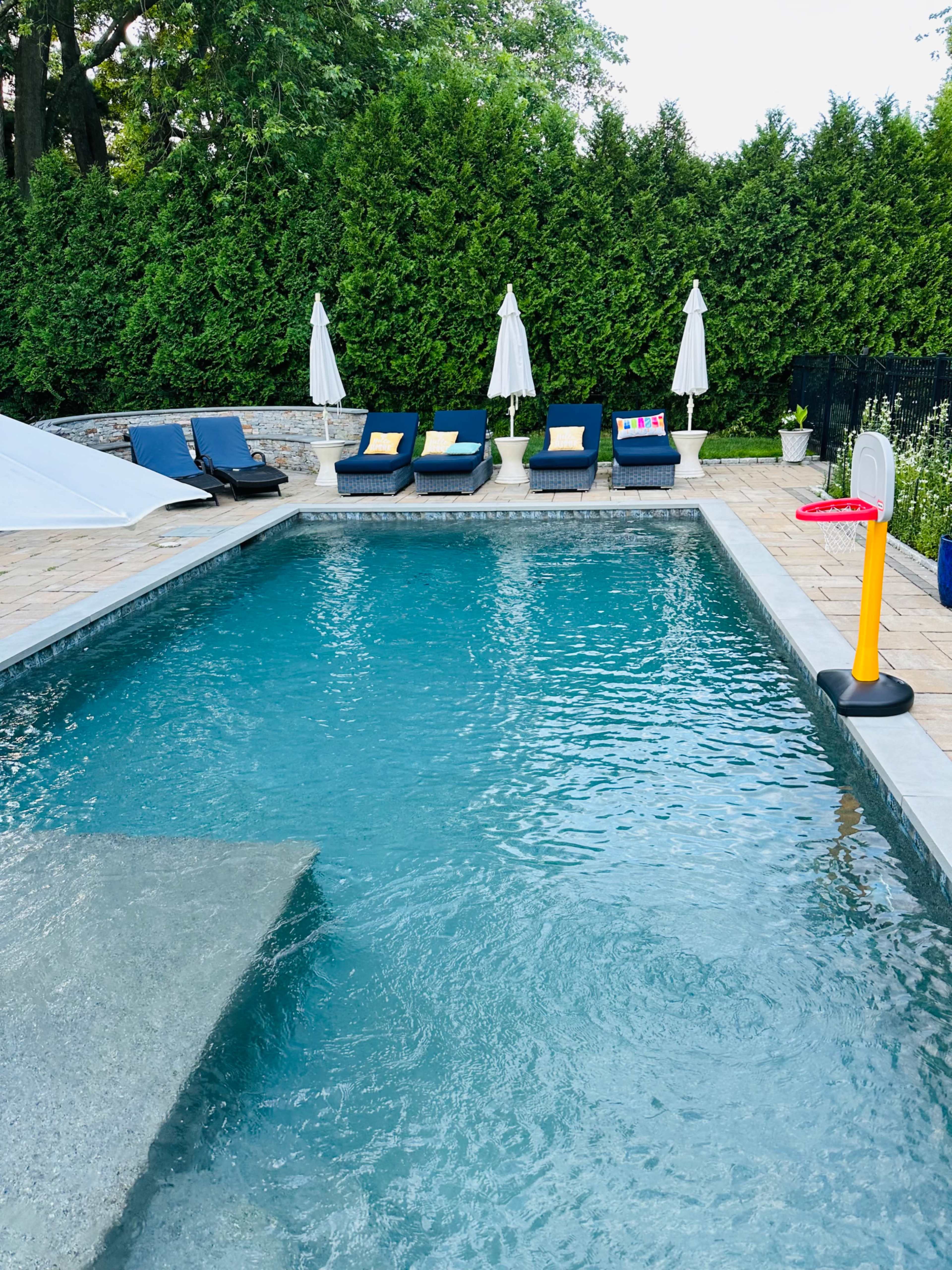 A clear swimming pool is surrounded by lounge chairs and umbrellas, with a basketball hoop nearby.