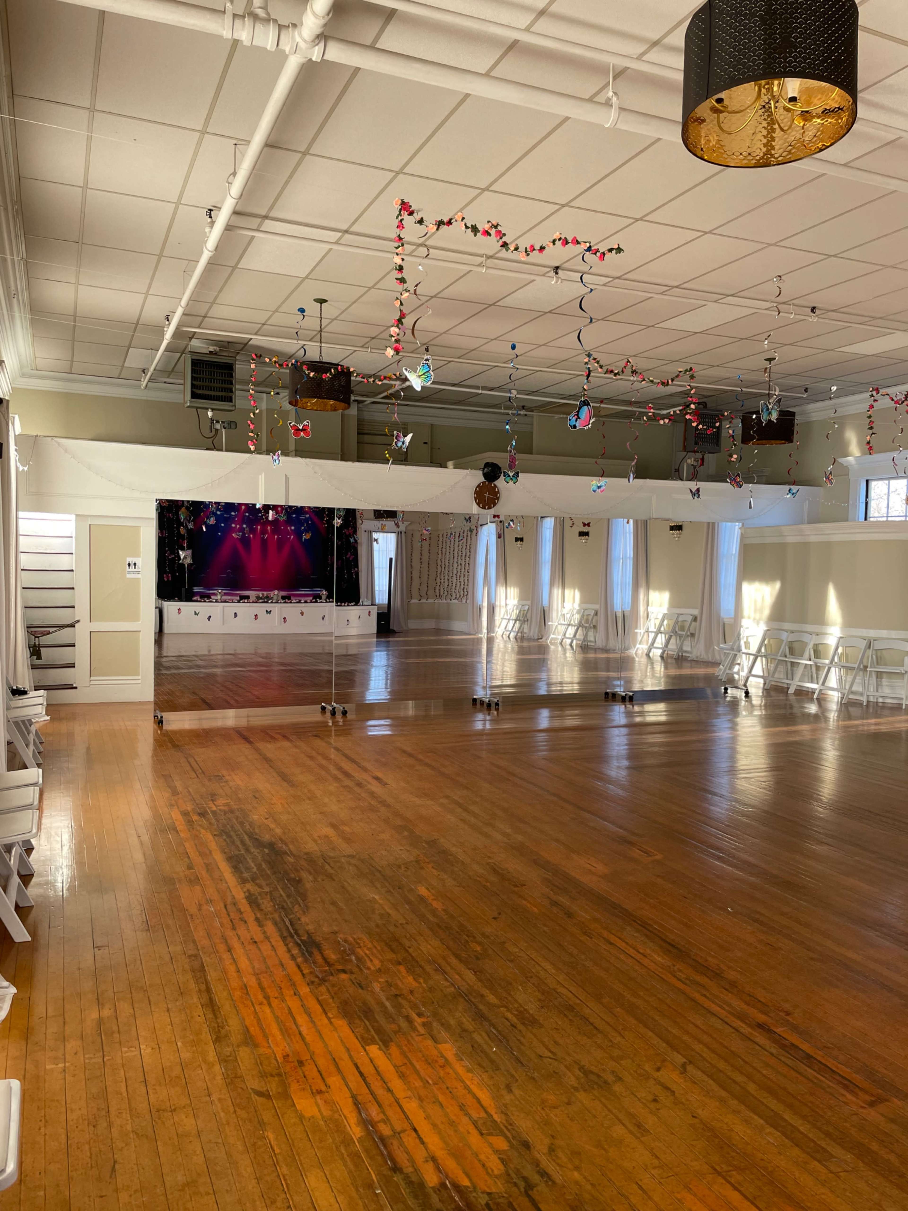 The image shows a spacious hall with a wooden floor, decorated with colorful streamers and lights, and a stage at one end.