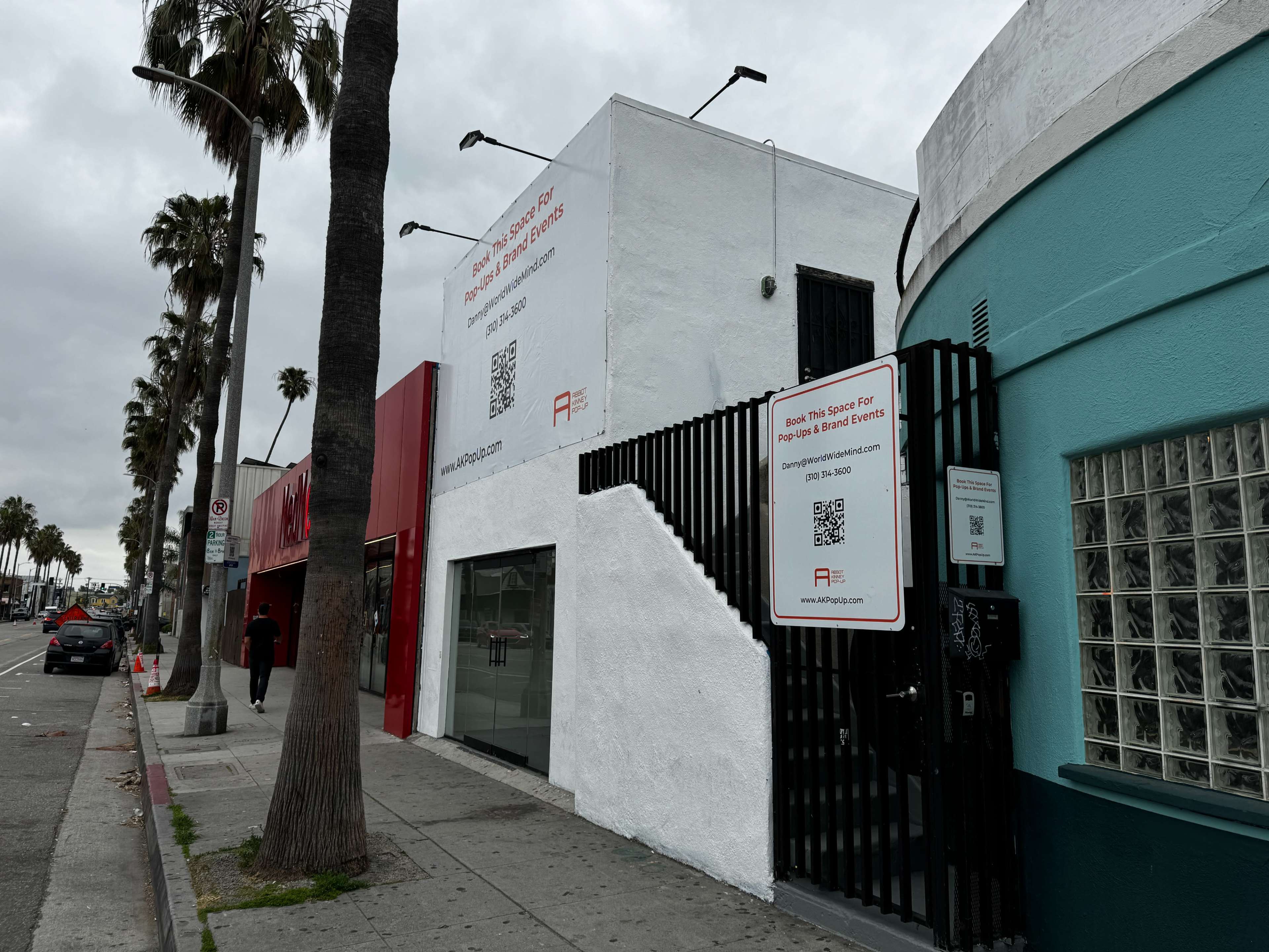 The image shows a white building with a large advertisement featuring QR codes and text, situated next to a teal building, along a street lined with palm trees.