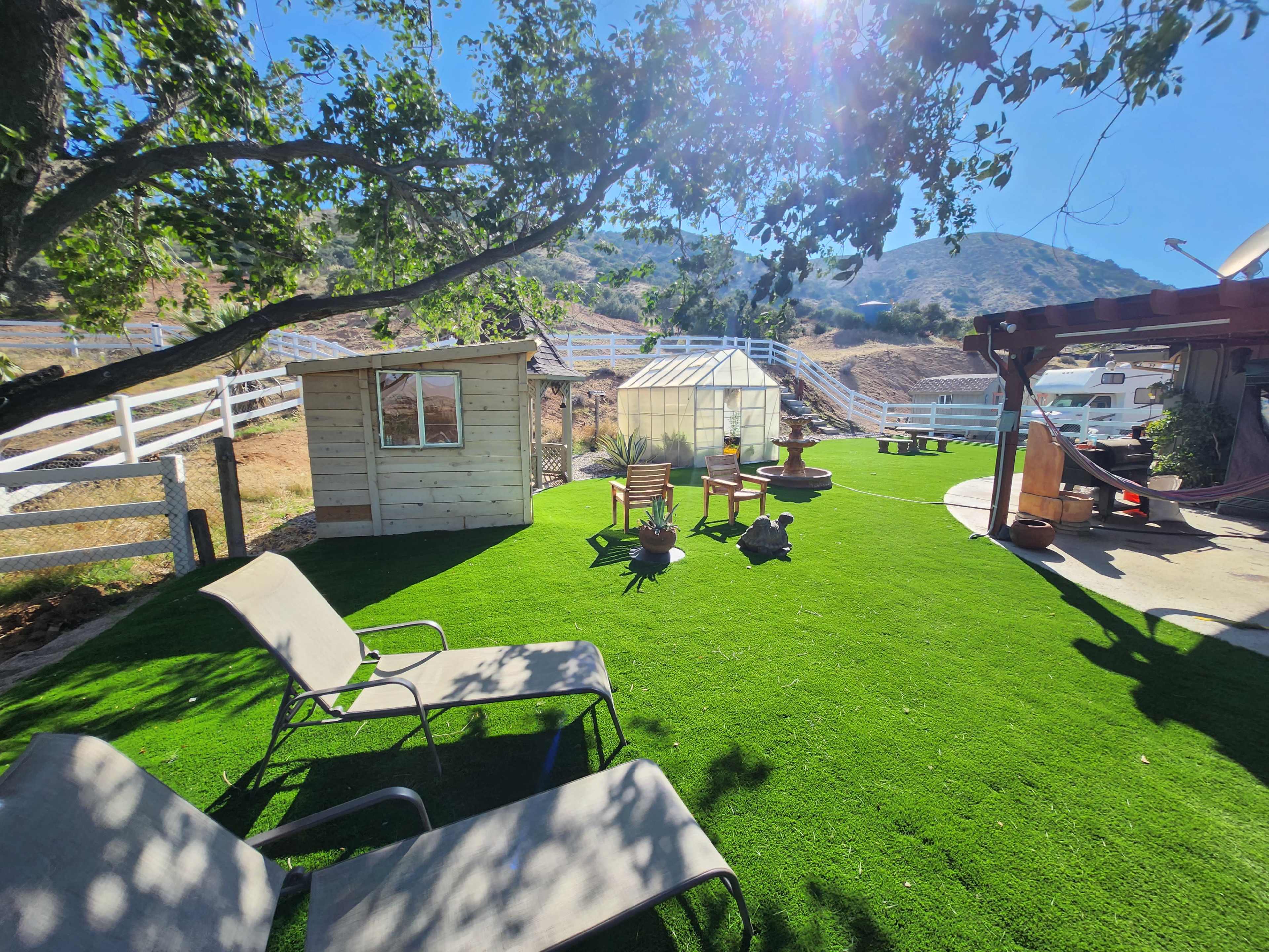 The scene shows a landscaped yard with artificial grass, a wooden shed, a greenhouse, and lounge chairs under a tree, with mountains in the background.