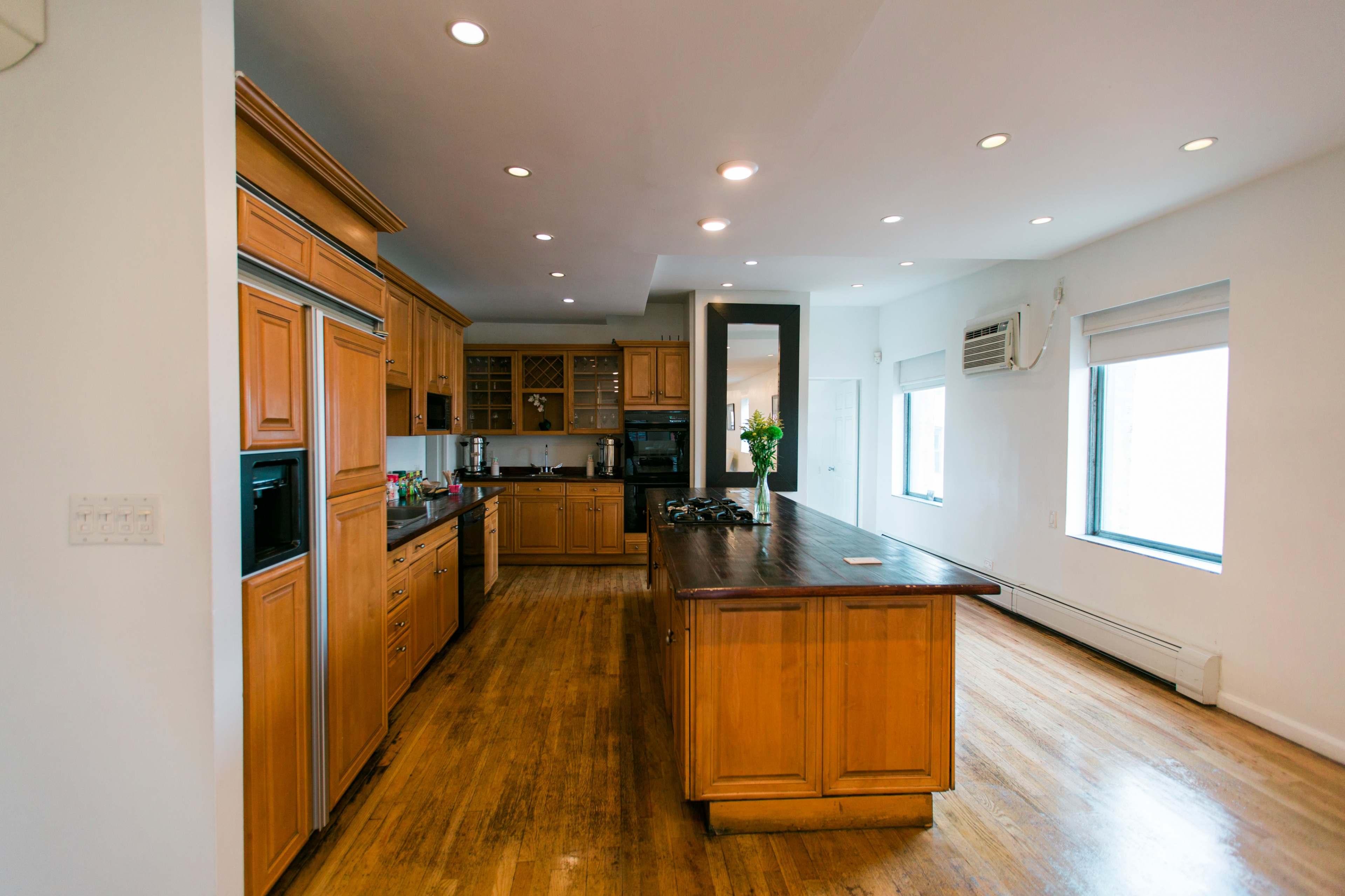 The image shows a spacious kitchen featuring wooden cabinets, a large island, and multiple windows for natural light.