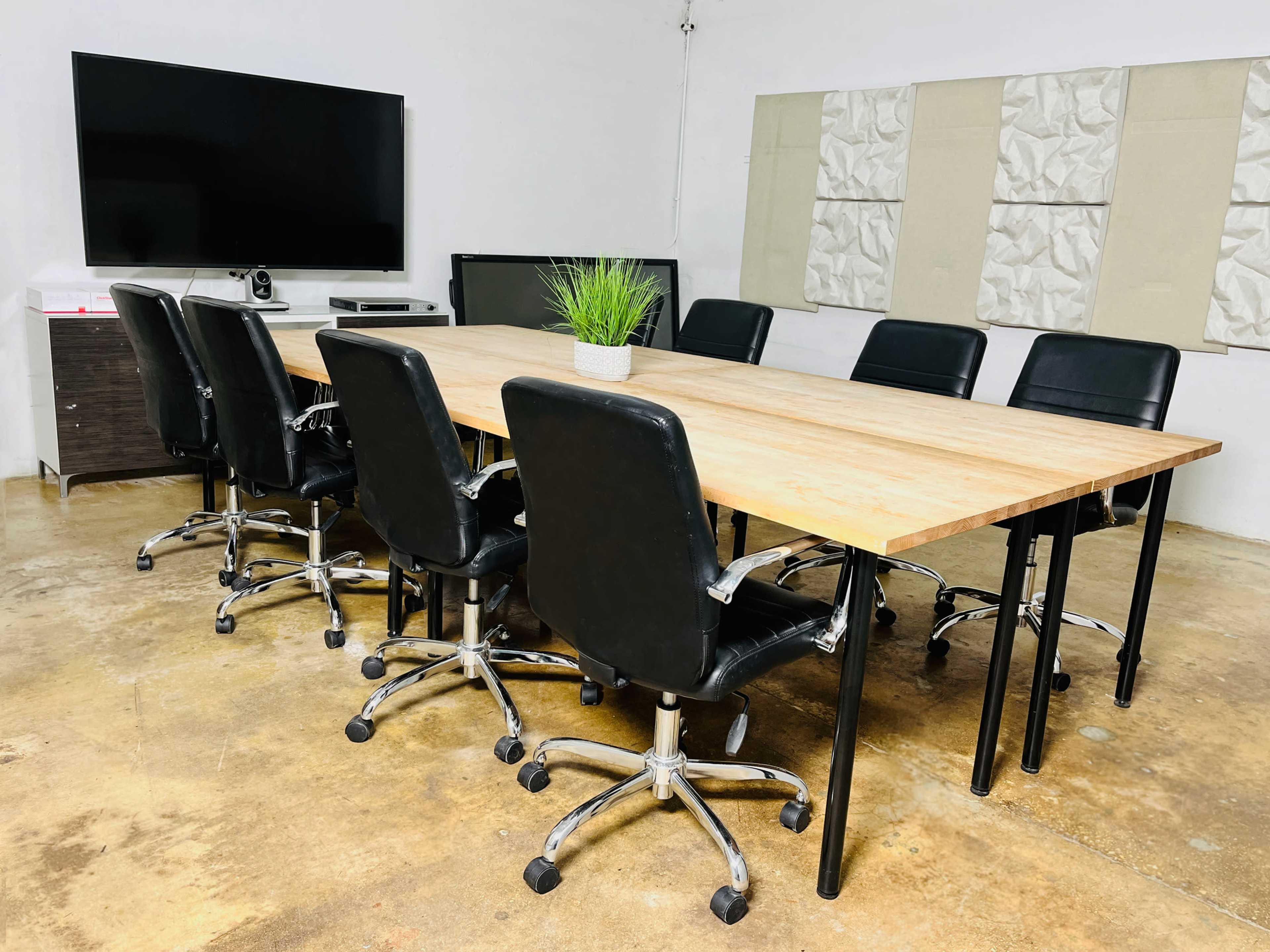 A conference room features a wooden table surrounded by seven black swivel chairs, with a television mounted on the wall and a small plant on the table.
