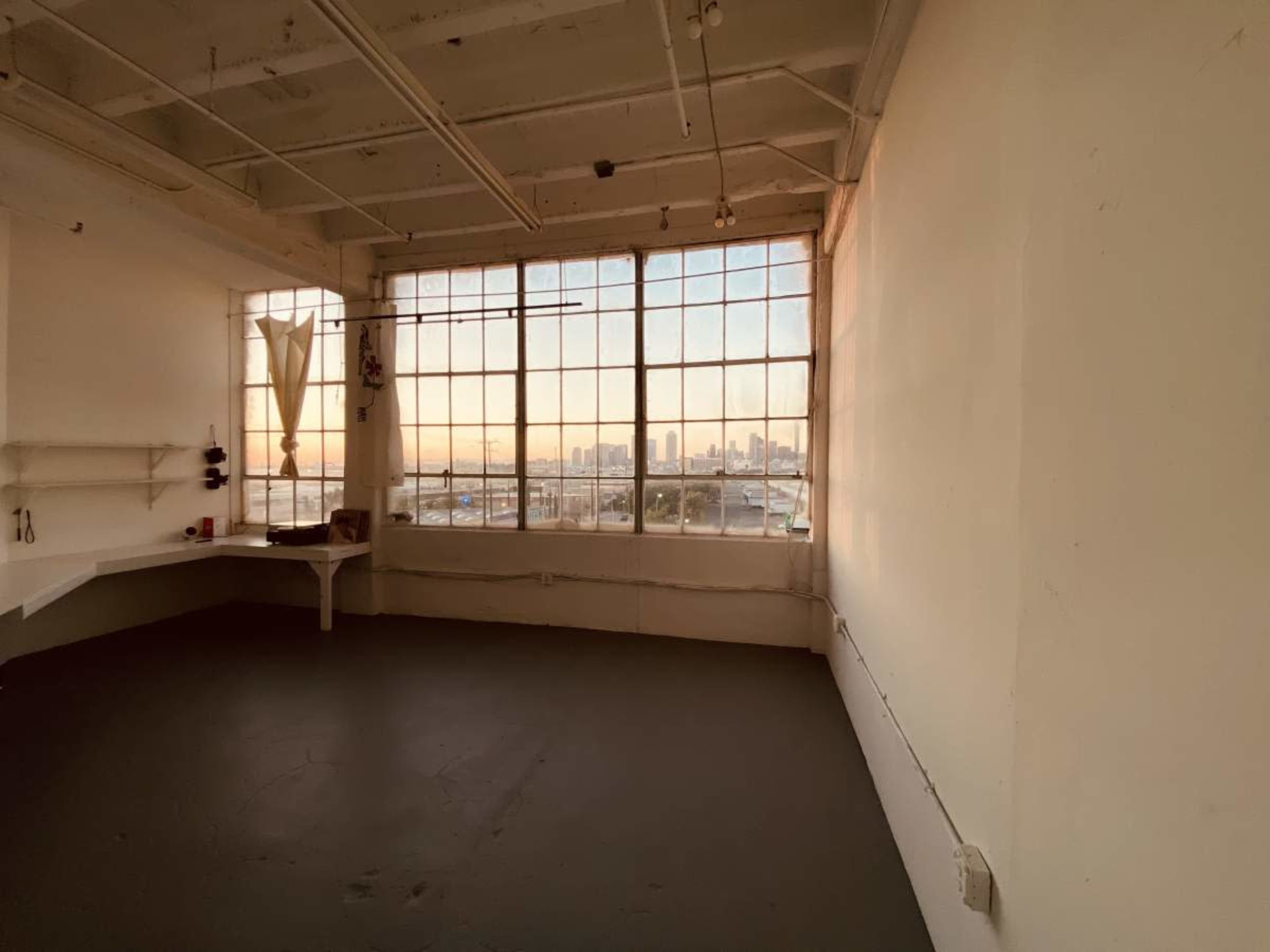 The image shows a spacious, empty room with concrete flooring and large windows that overlook a city skyline at sunset.