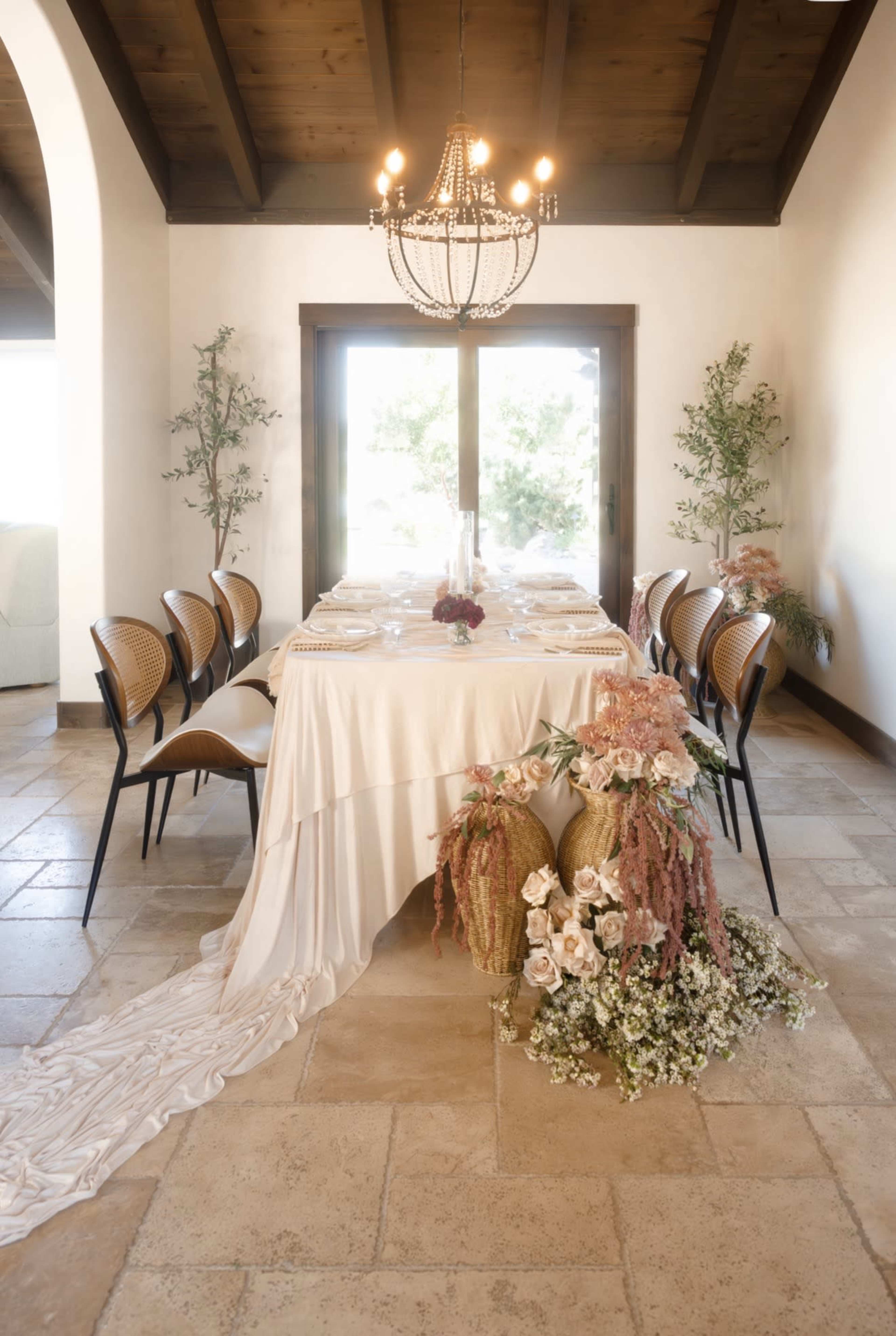 A long dining table is decorated with a white tablecloth, surrounded by chairs, and adorned with floral arrangements and greenery.