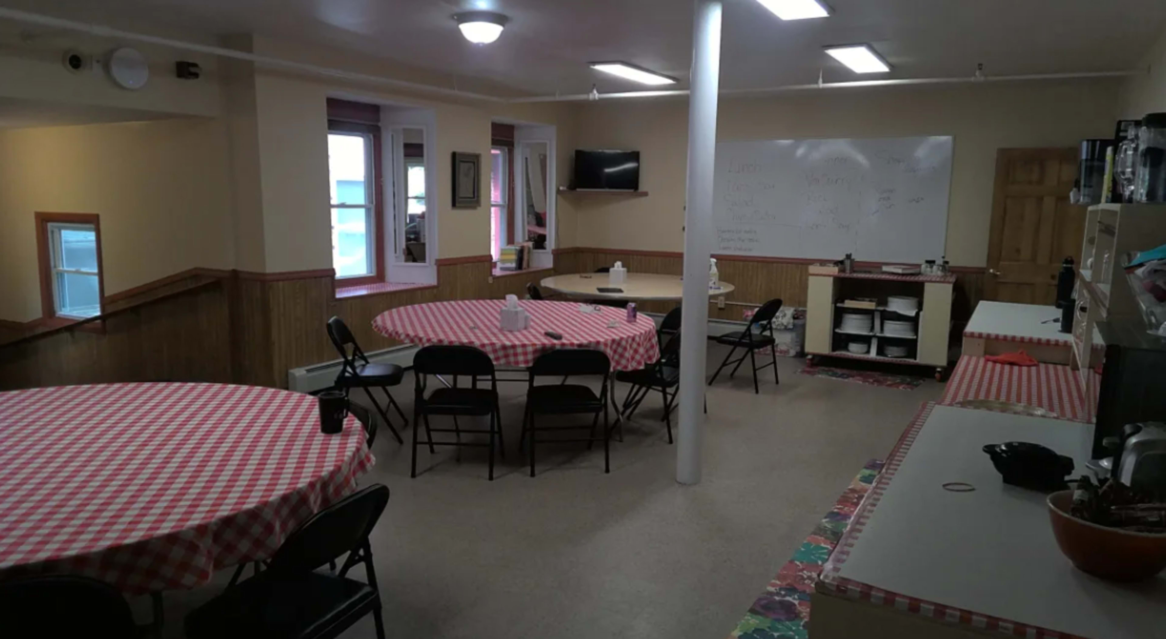 A spacious indoor dining area with several round tables covered in red and white checkered tablecloths, a kitchenette along one wall, and a whiteboard on another.