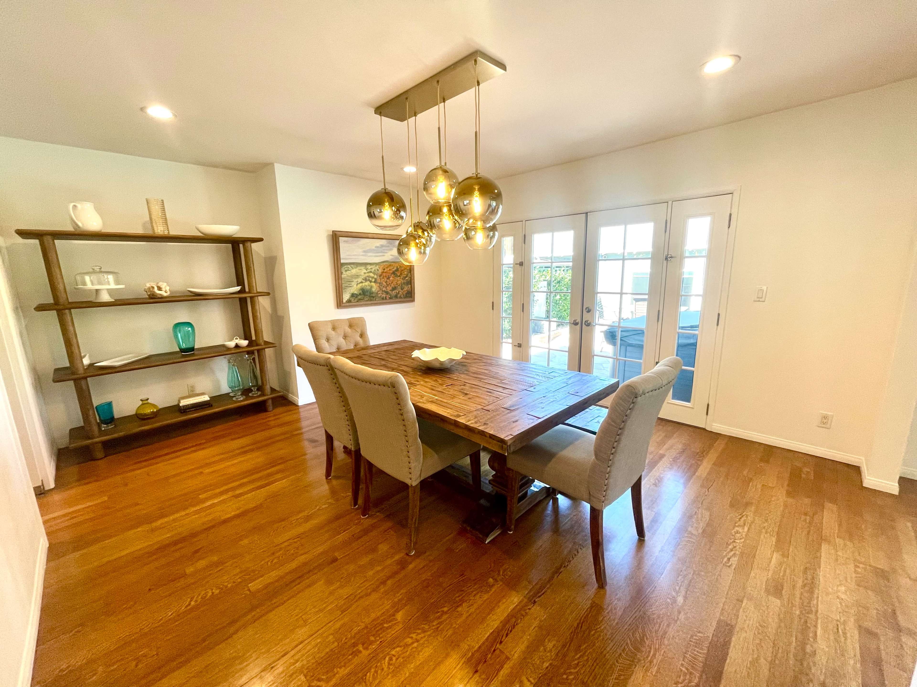 A dining room features a wooden table with upholstered chairs, a shelf displaying decorative items, and a light fixture above.