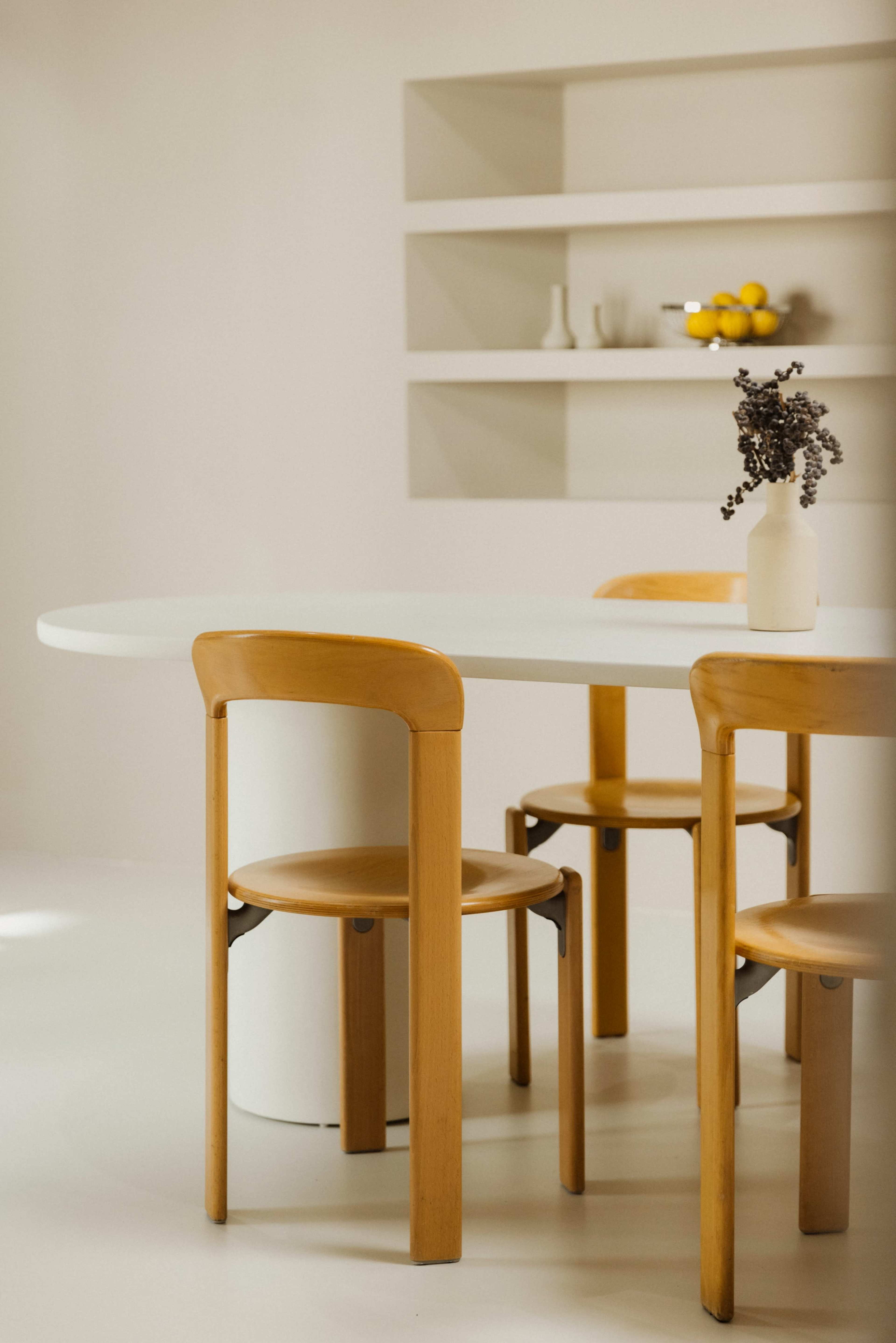 A minimalist dining area features a round white table with three light wood chairs and a vase of dried flowers, against a pale background with a shelf displaying lemons and decorative items.