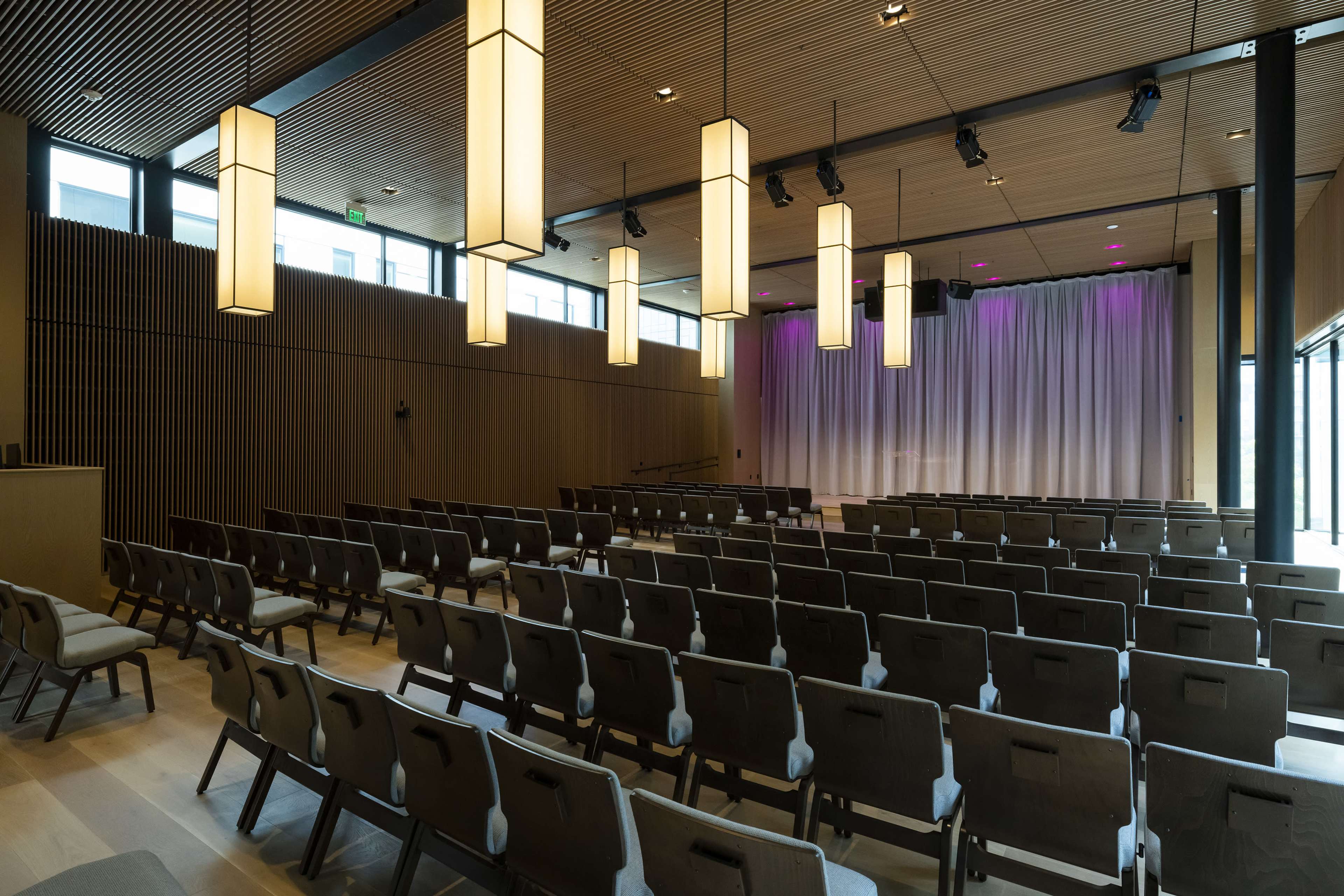 A modern auditorium with rows of chairs facing a stage, accented by hanging light fixtures and large windows.