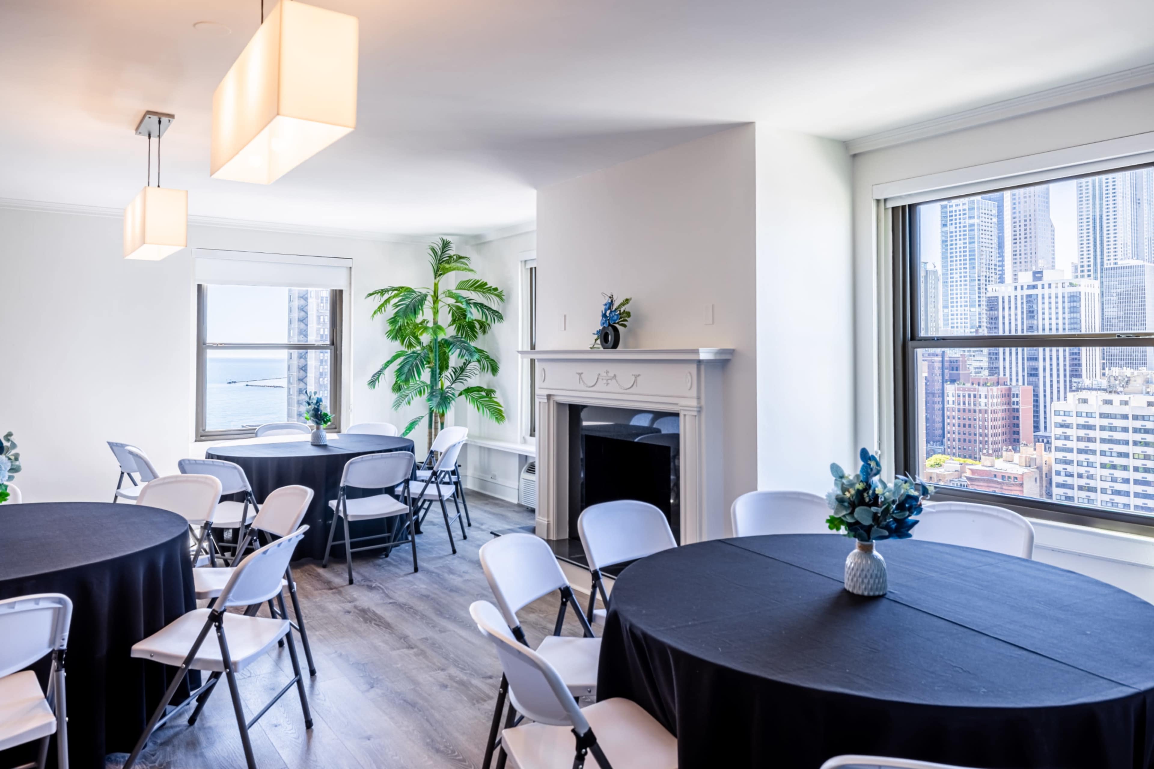 The image shows a bright, modern event space featuring round tables with black tablecloths, white chairs, and large windows offering a view of a cityscape.