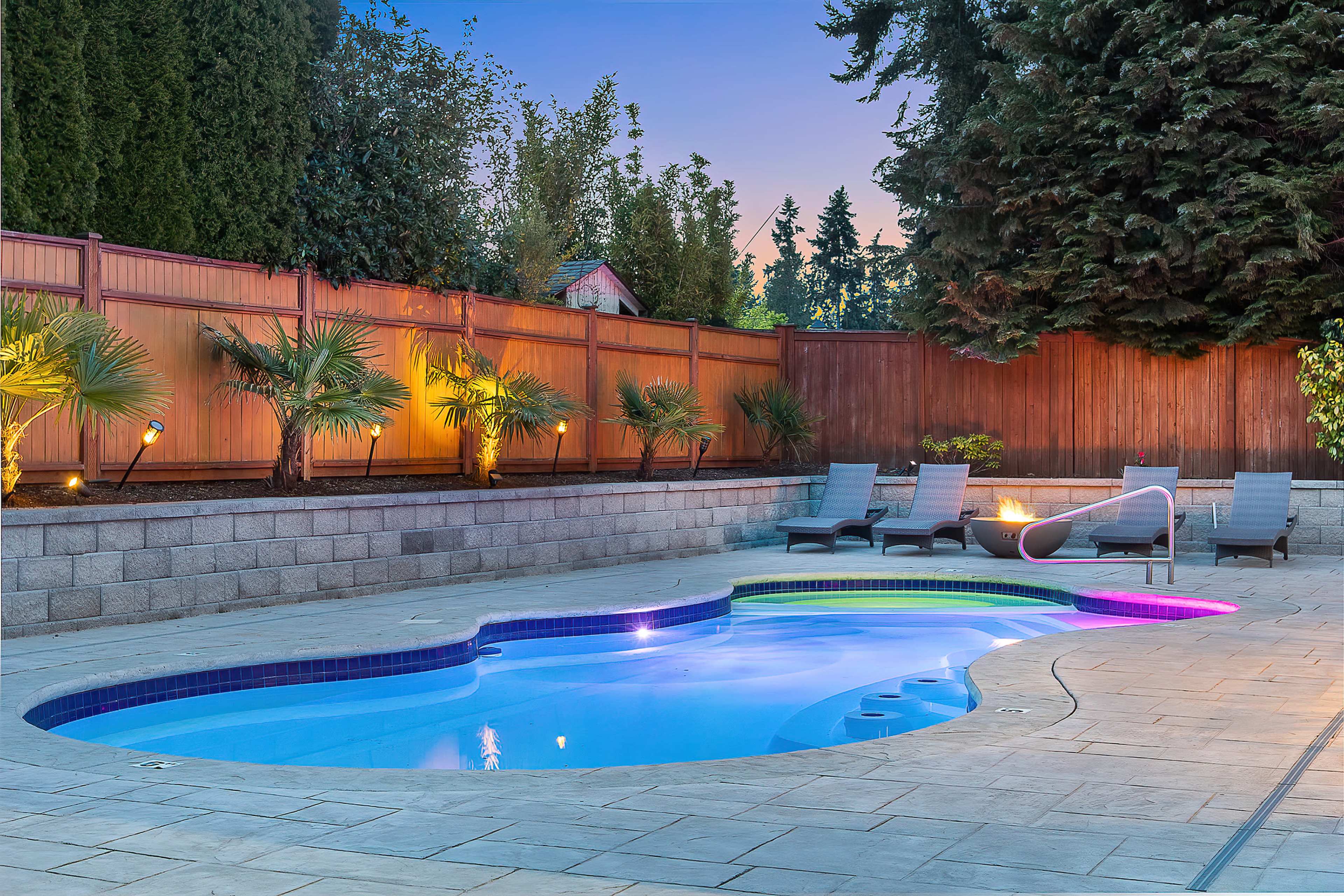 The image shows a well-lit outdoor pool area with lounge chairs and palm trees surrounded by a wooden fence.
