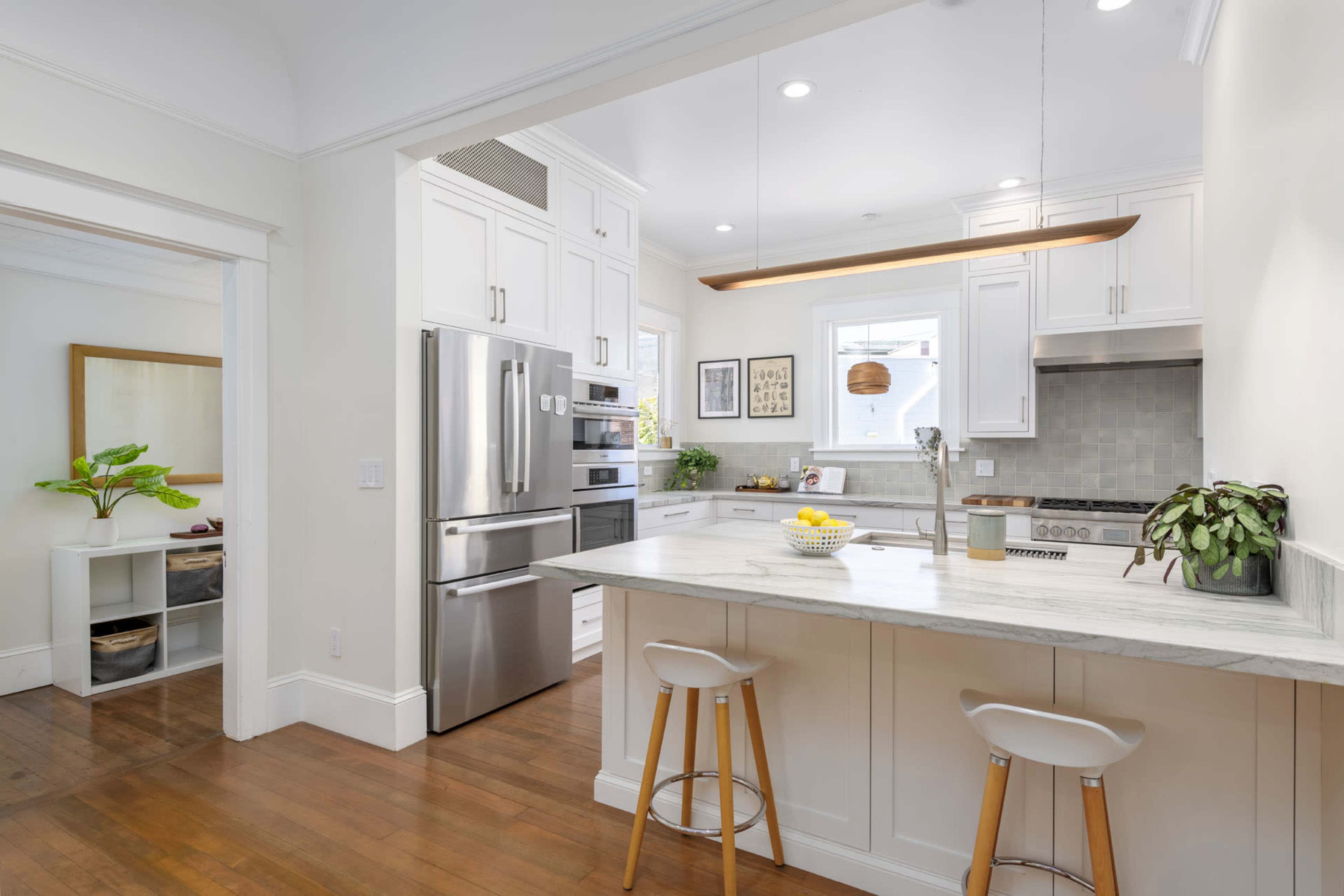 A modern kitchen features white cabinetry, stainless steel appliances, and a central island with bar stools, set against warm wooden flooring.