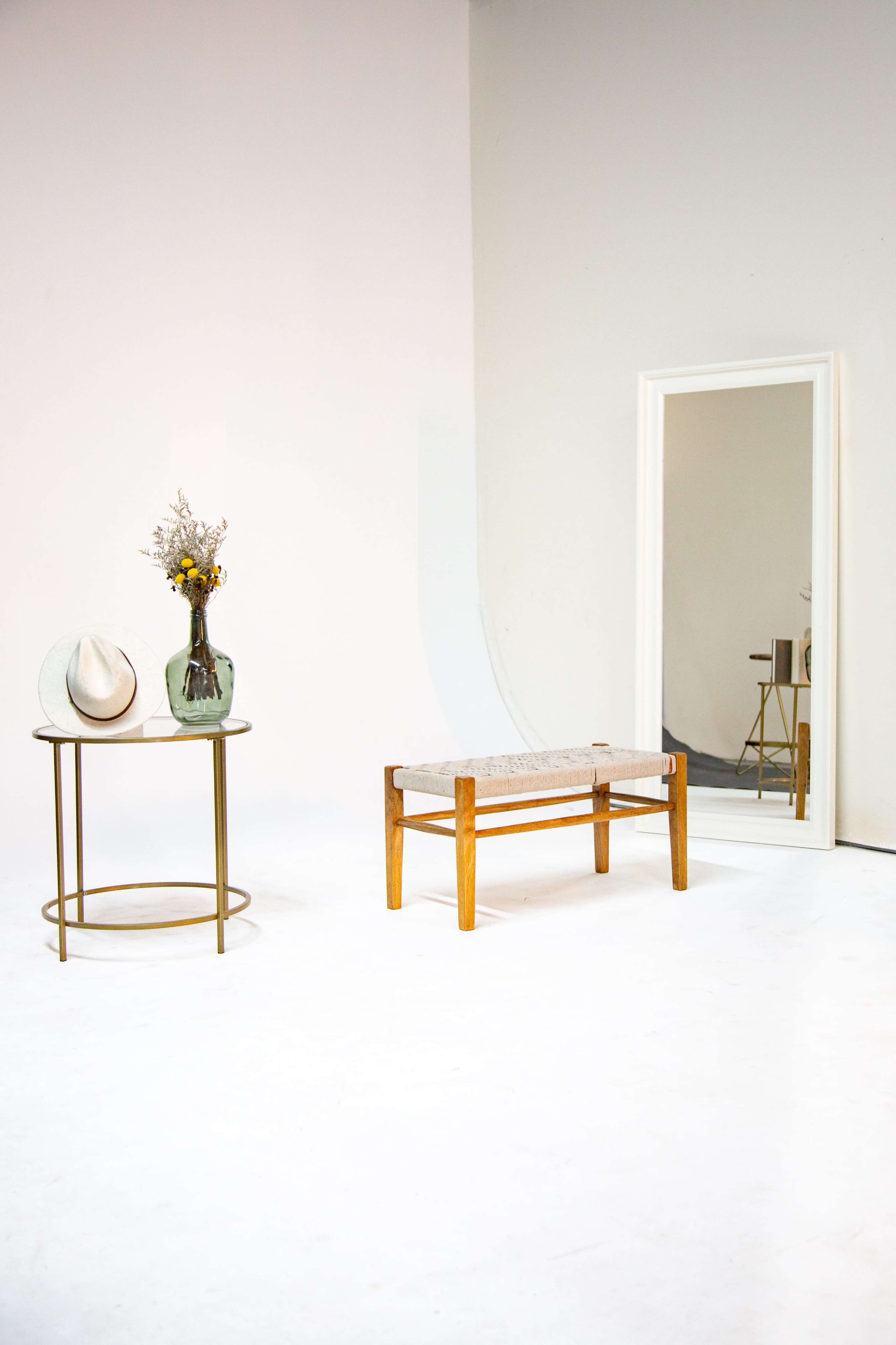 The scene features a minimalist interior with a round table holding a vase of flowers and a straw hat, alongside a wooden bench and a large mirror against a light-colored backdrop.