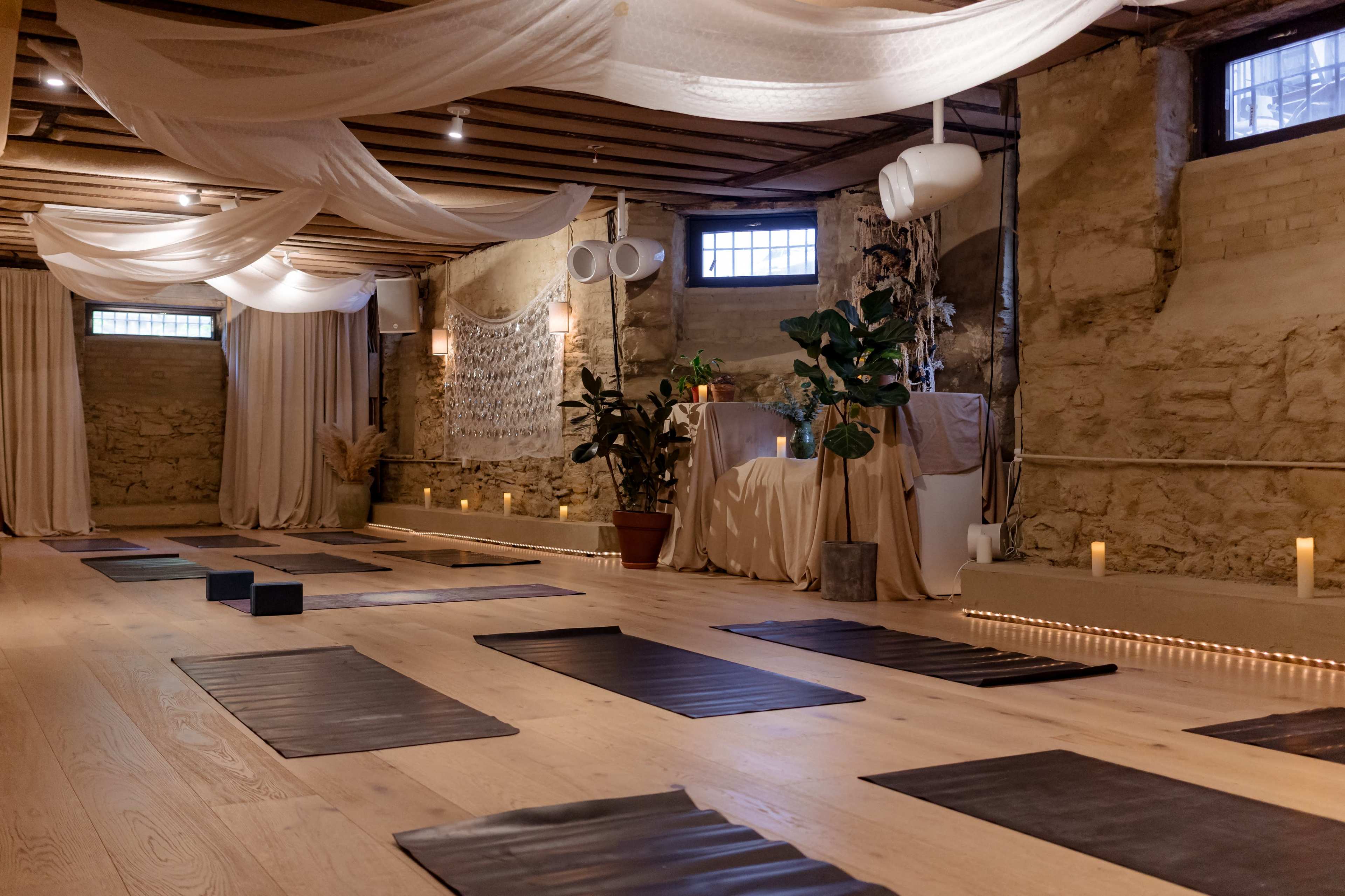 The image shows a serene yoga studio with wooden flooring, yoga mats neatly arranged, and soft lighting illuminating the space.