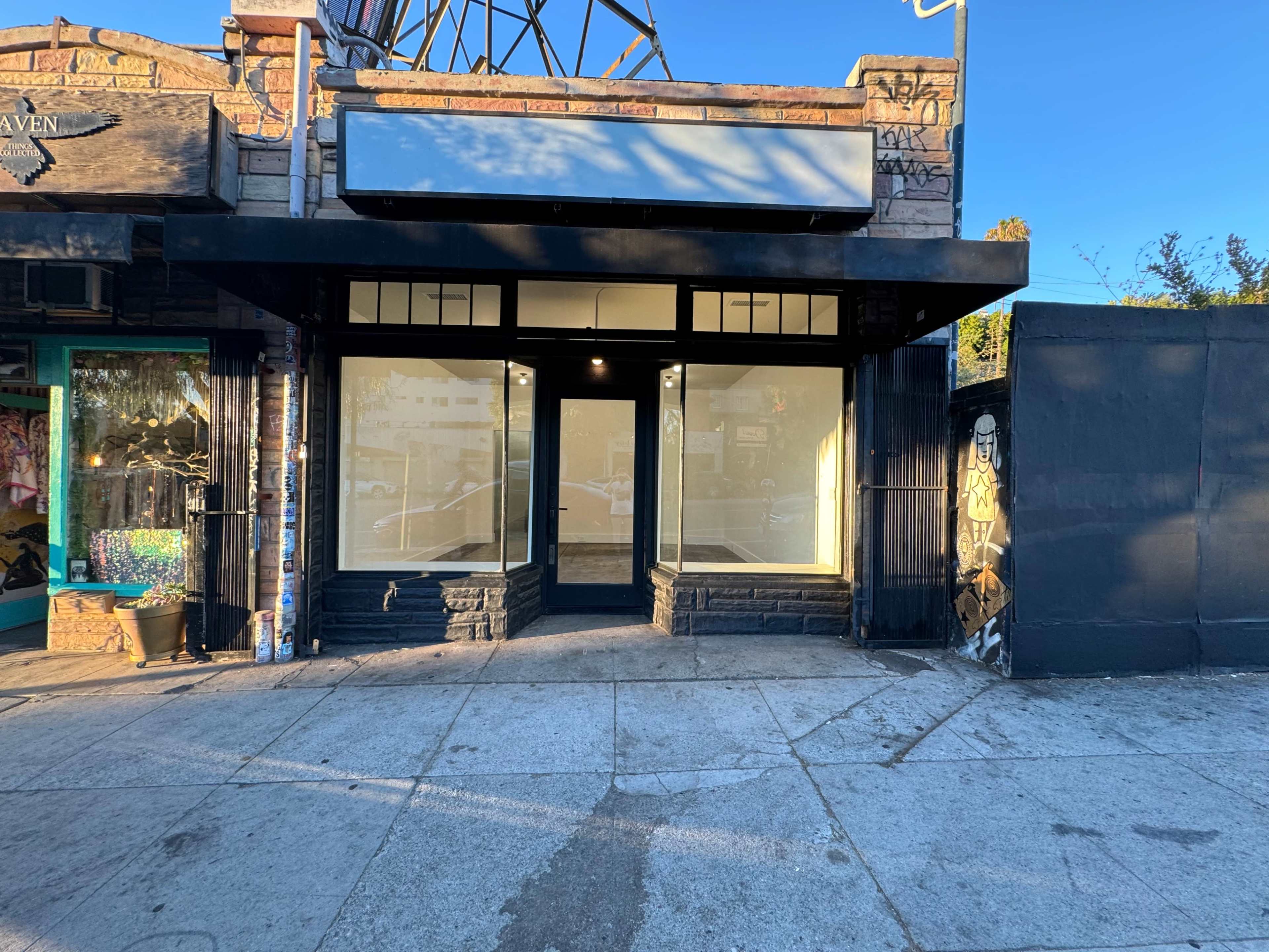 The image shows a vacant retail storefront with large windows and minimal signage, located on a sidewalk beside a painted wall.