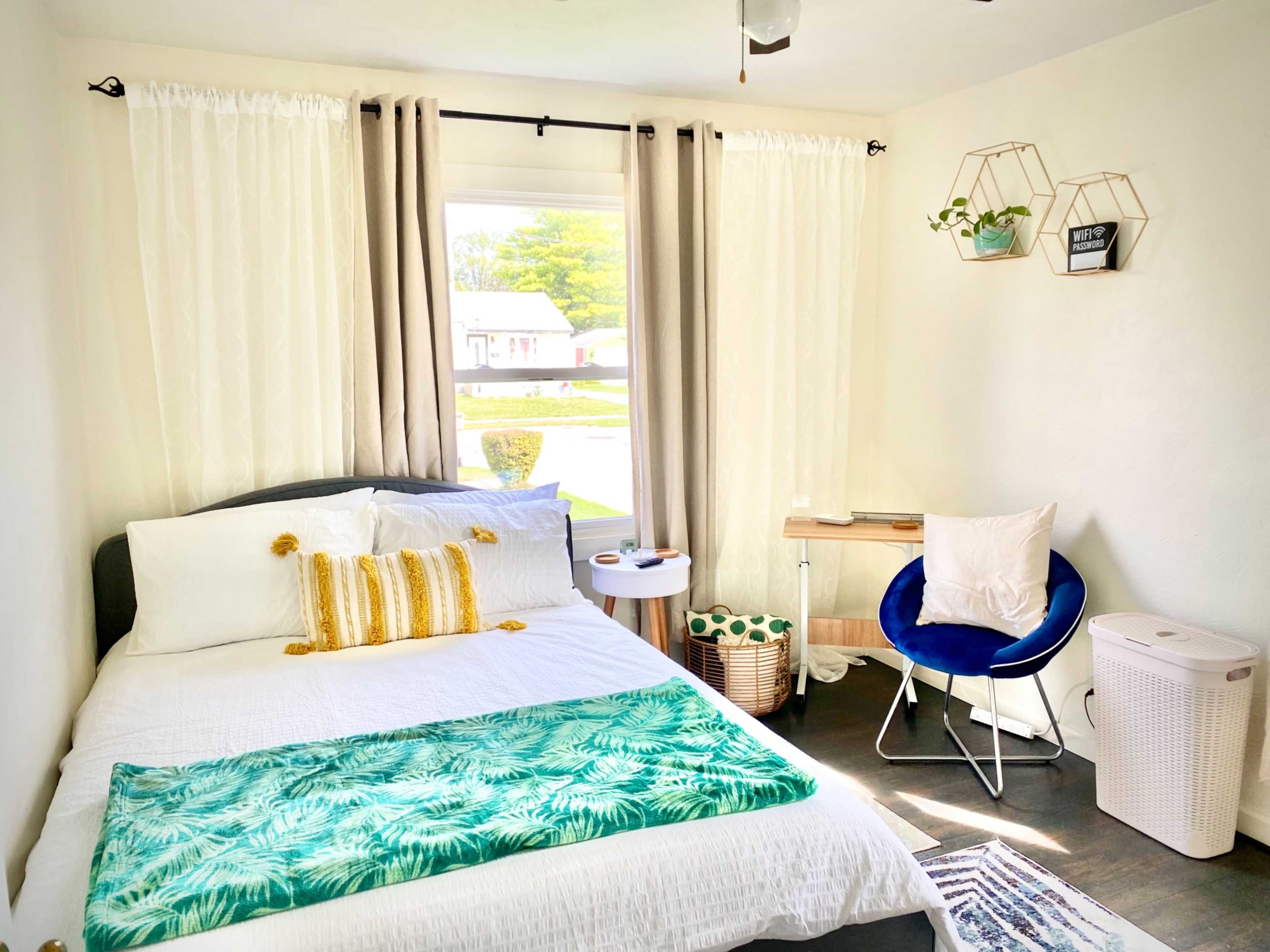 A cozy bedroom featuring a double bed with white linens, a small table, a blue chair, and large windows with light curtains.
