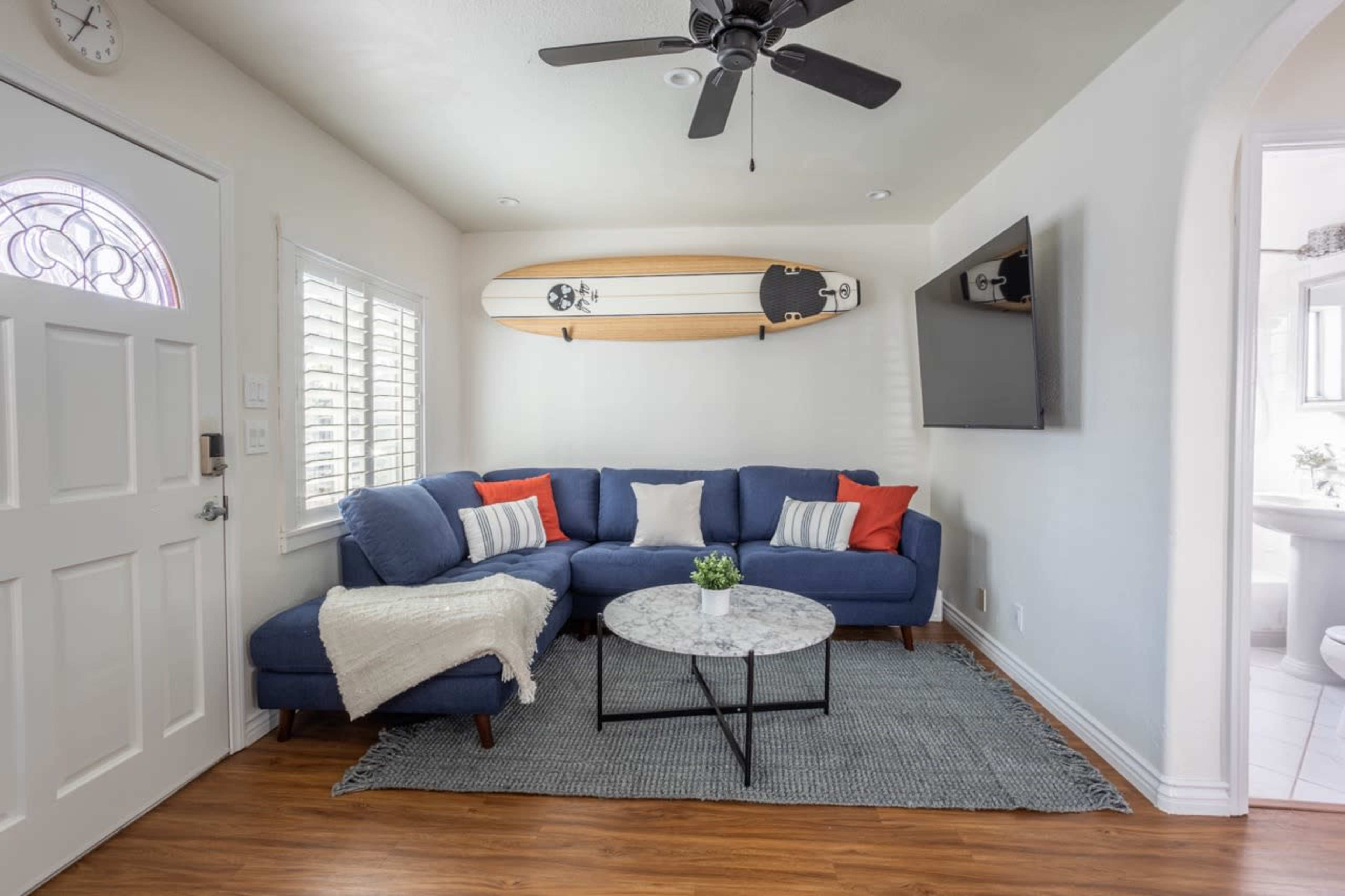 A modern living room features a blue sectional sofa with red and white cushions, a round coffee table, and a surfboard mounted on the wall.