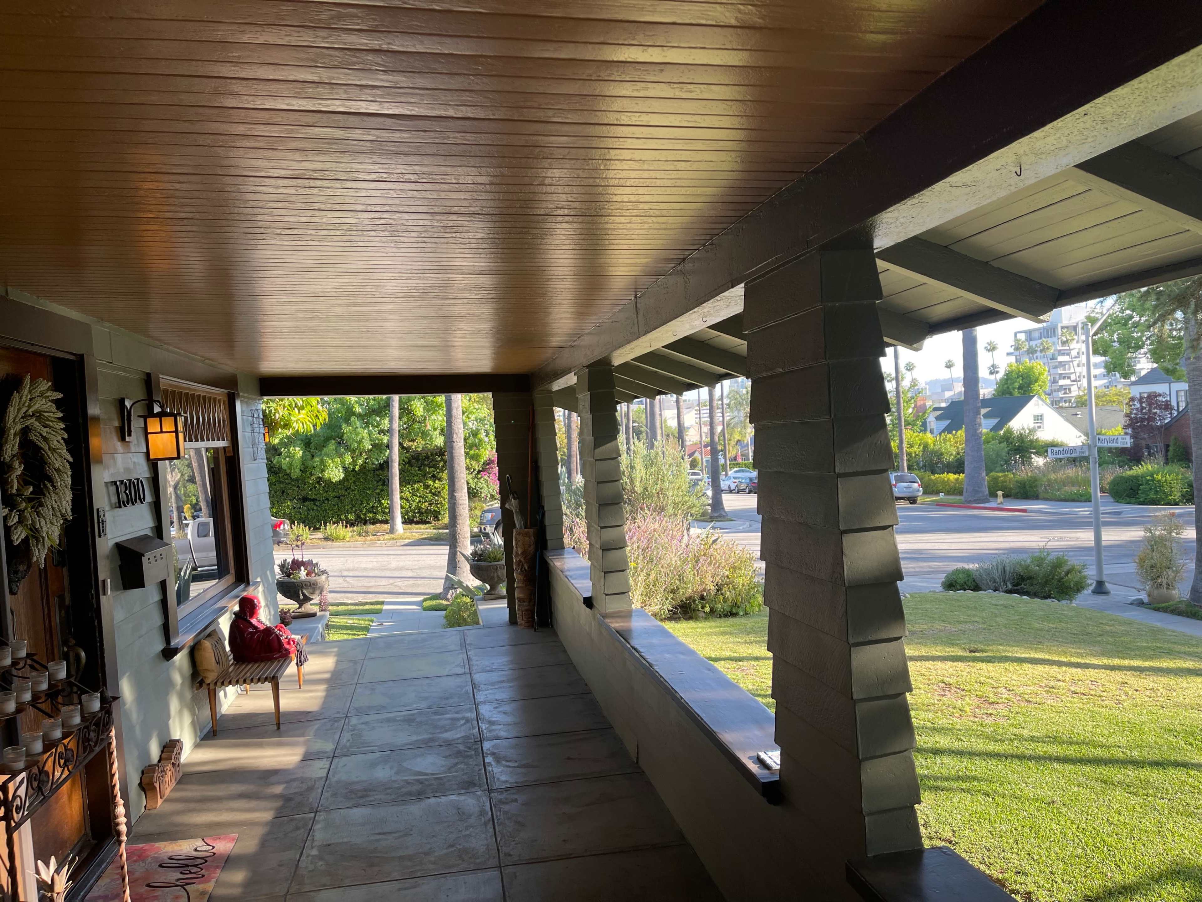 The image shows a covered porch with a bench and decorative elements, facing a street lined with trees and greenery.