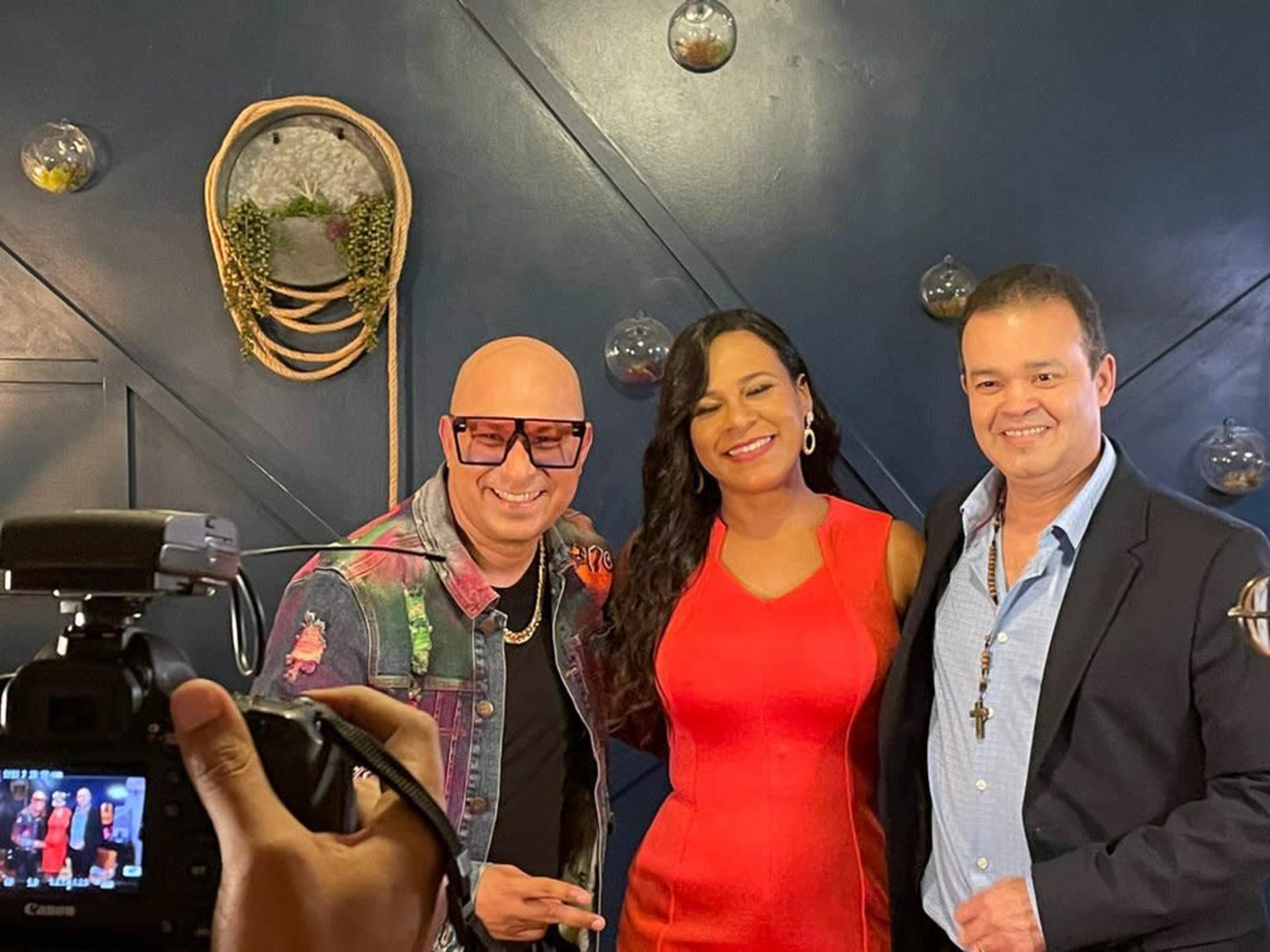 Three individuals pose for a photo in front of a dark blue wall decorated with hanging planters and glass orbs.