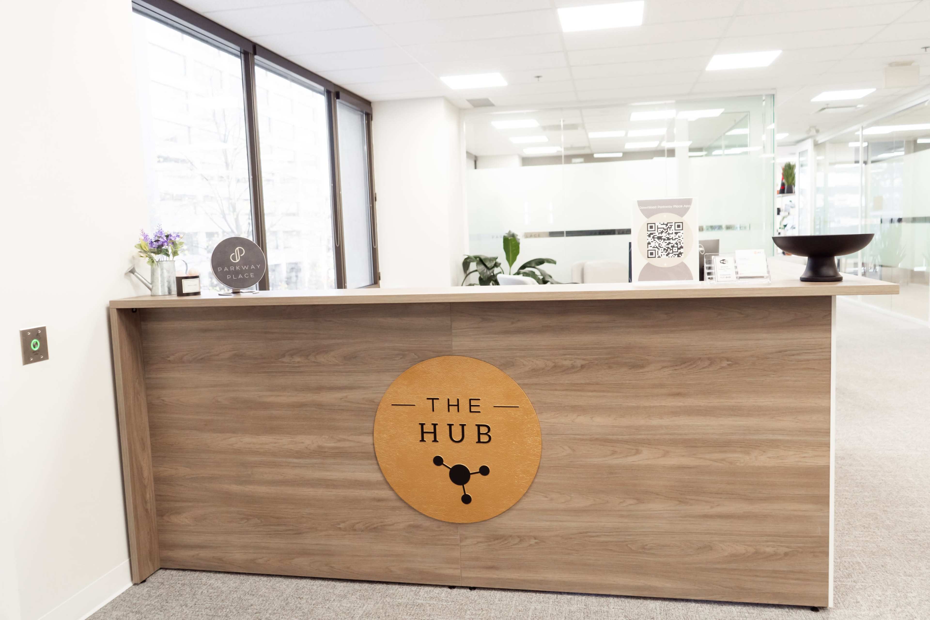 The image shows a modern reception desk labeled "THE HUB" in a bright office space with large windows.