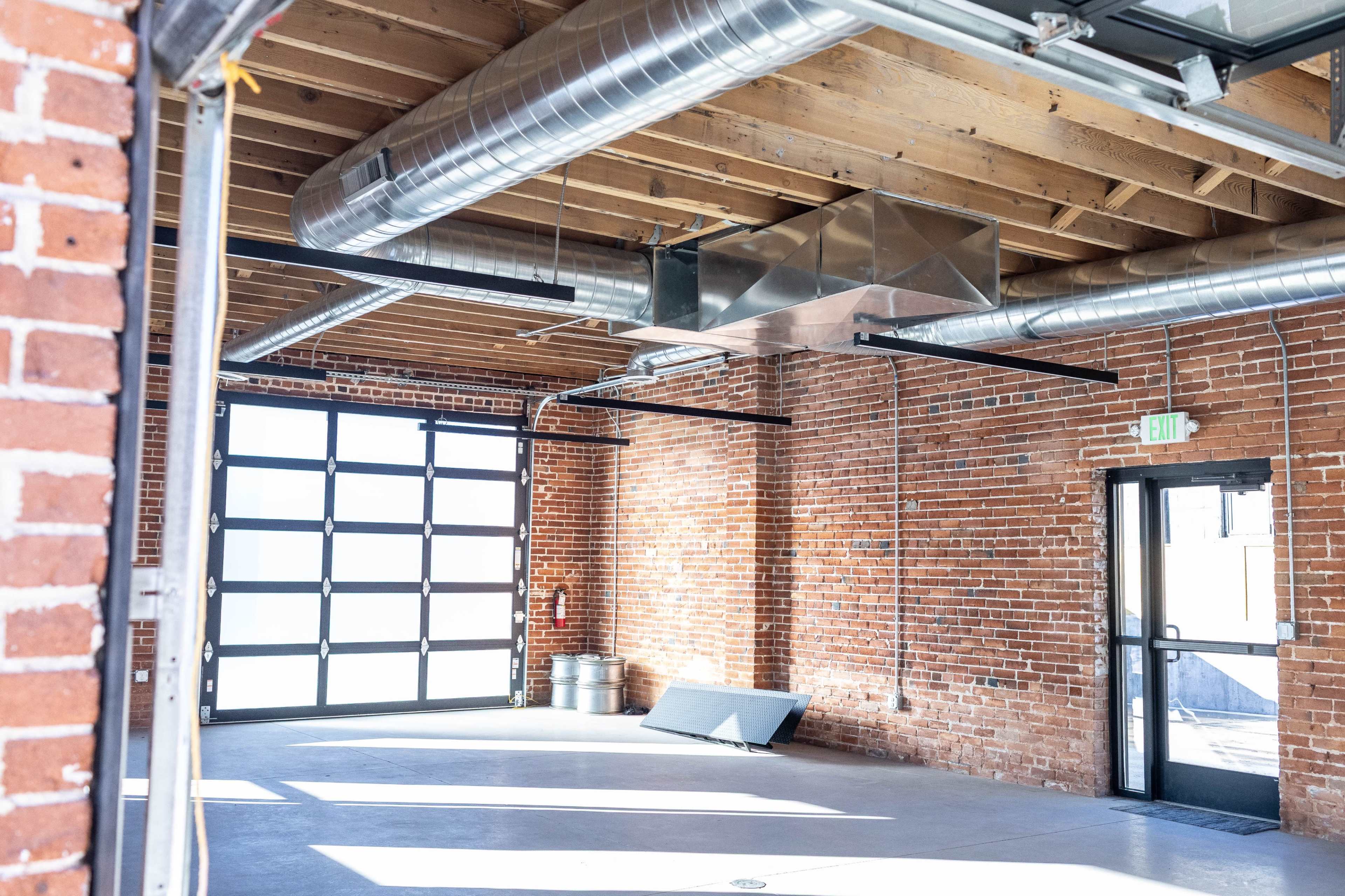 The image shows an industrial-style interior space with exposed brick walls, metal ductwork, and a large garage door.