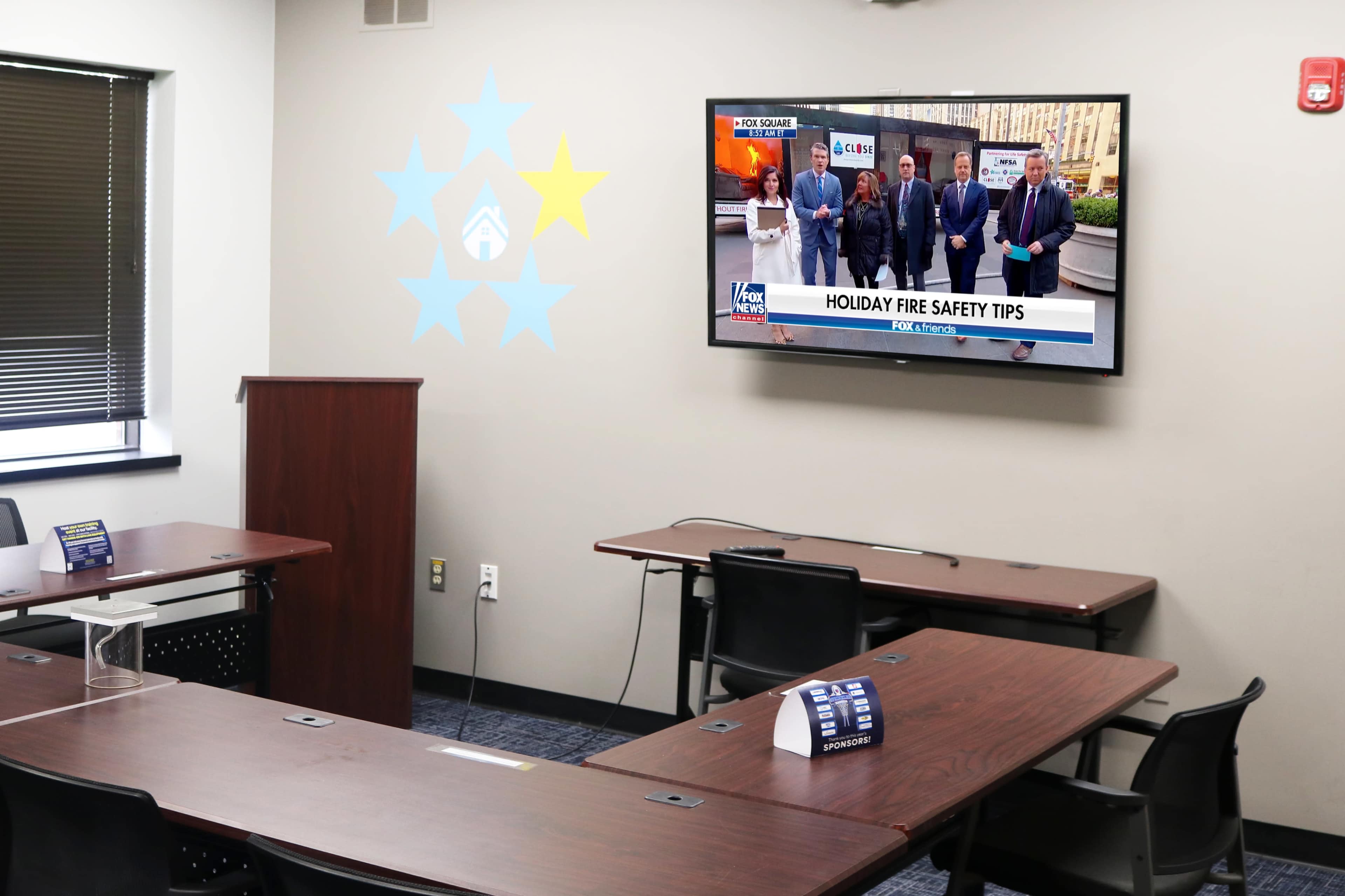 A meeting room is set up with tables and chairs, featuring a wall-mounted television displaying a segment on holiday fire safety tips.