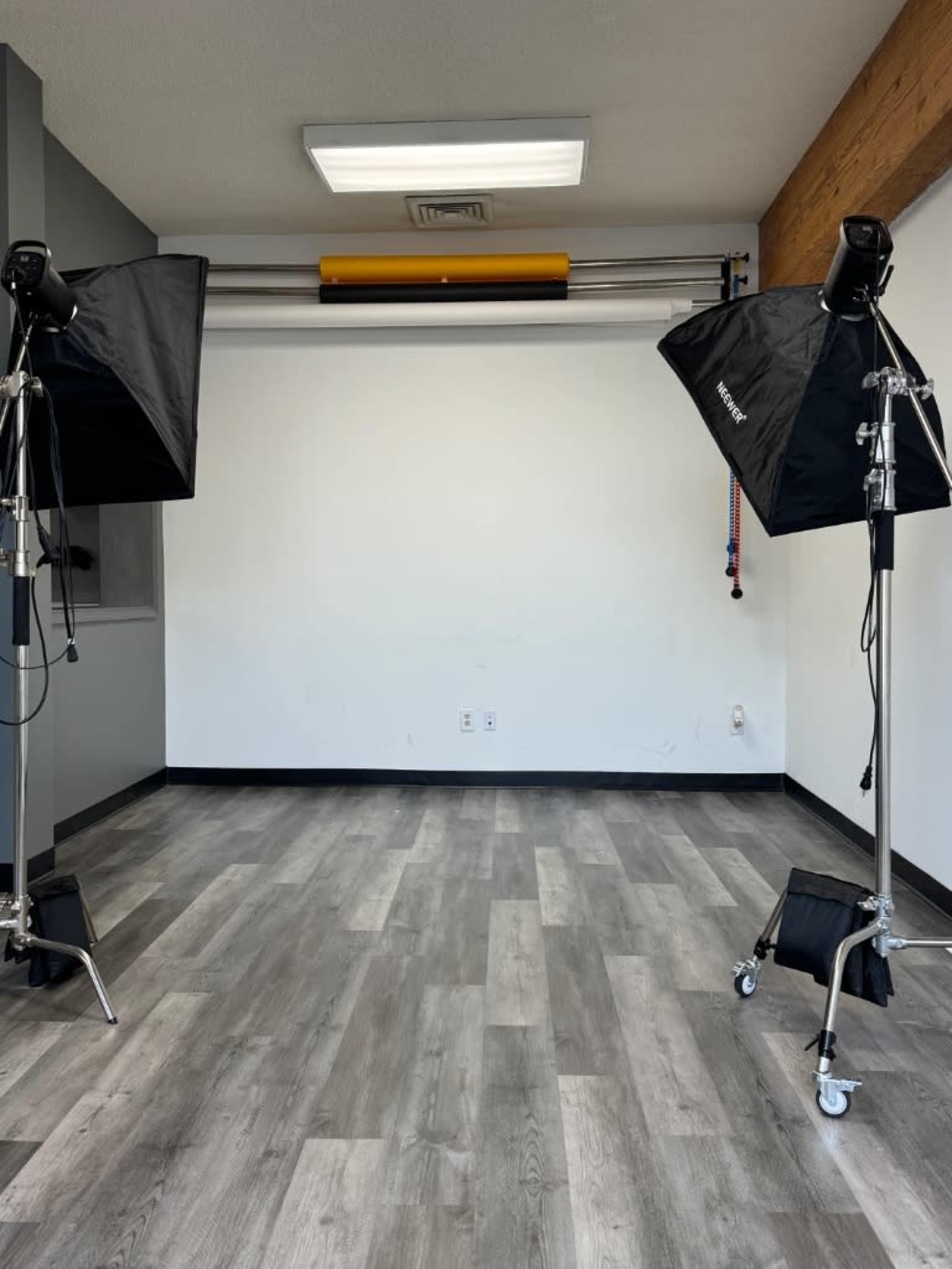 The image shows a small, minimally furnished studio space with two light stands positioned on either side of a blank wall.