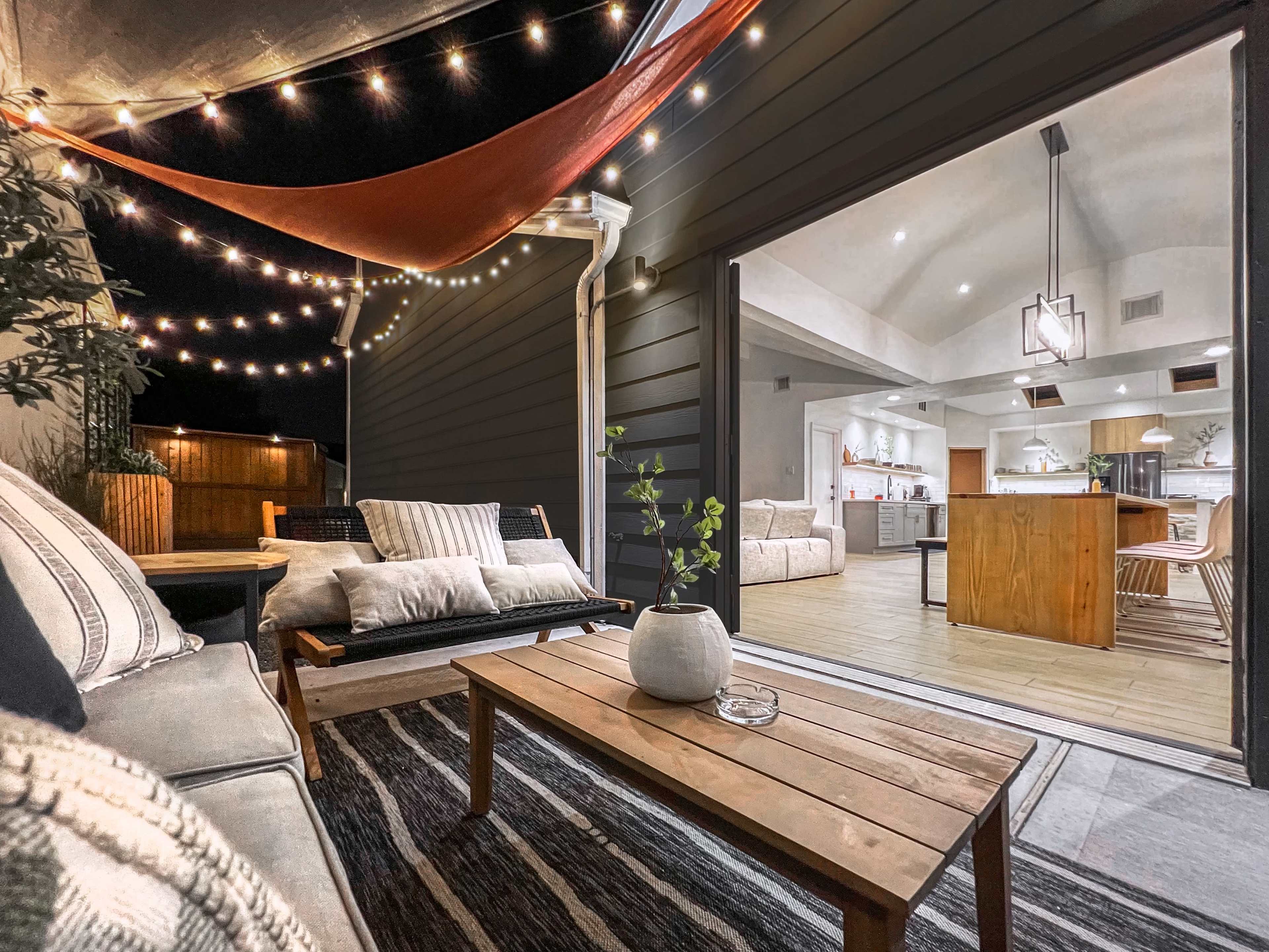 A cozy outdoor seating area illuminated by string lights, leading to a modern interior space with a kitchen visible through an open doorway.