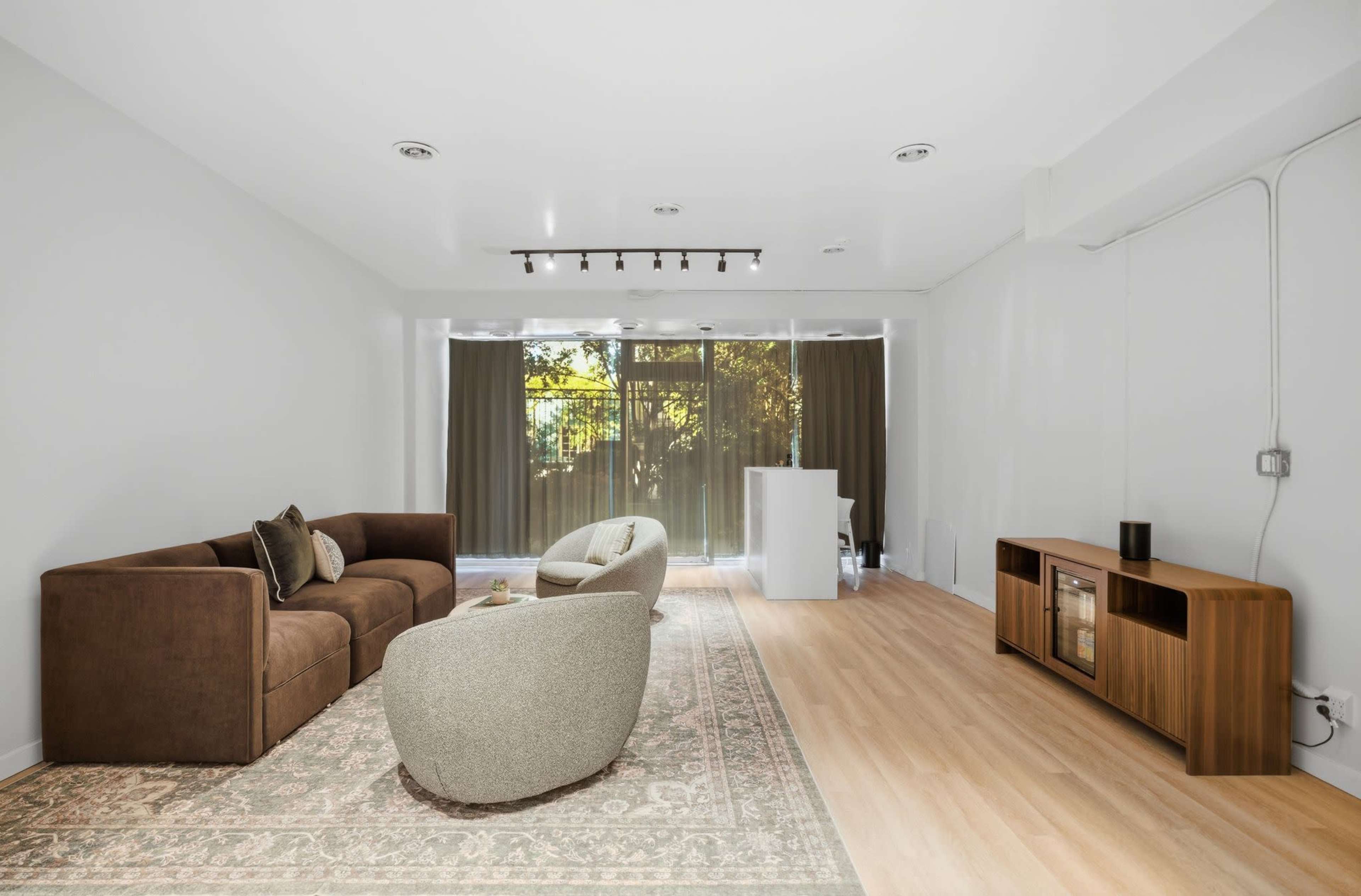 The image shows a spacious living room with a brown couch, a round beige chair, a wooden media console, and large windows covered by sheer curtains allowing natural light to enter.