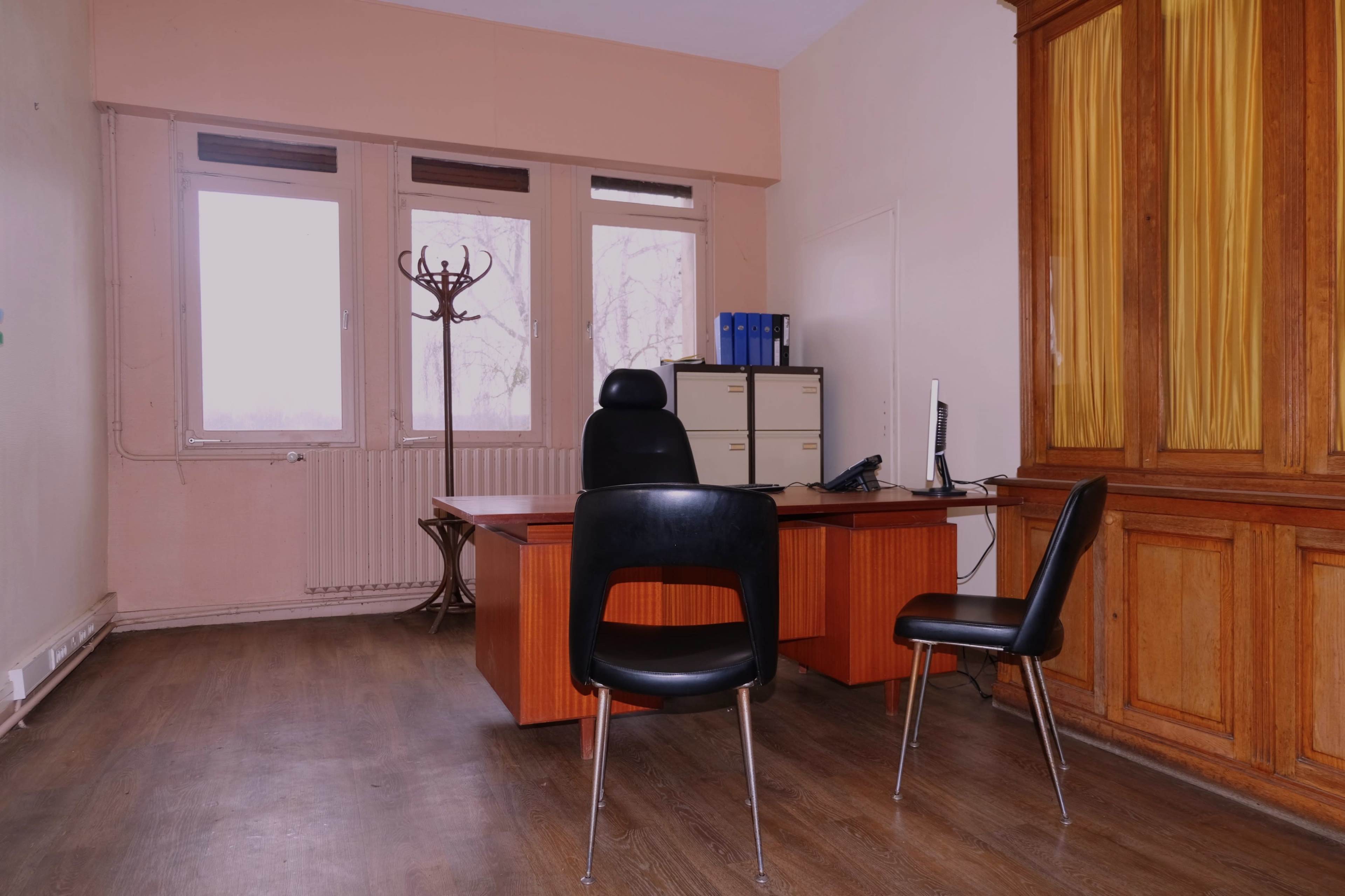 An empty office features a wooden desk, two chairs, a computer, and large windows overlooking a bare landscape.