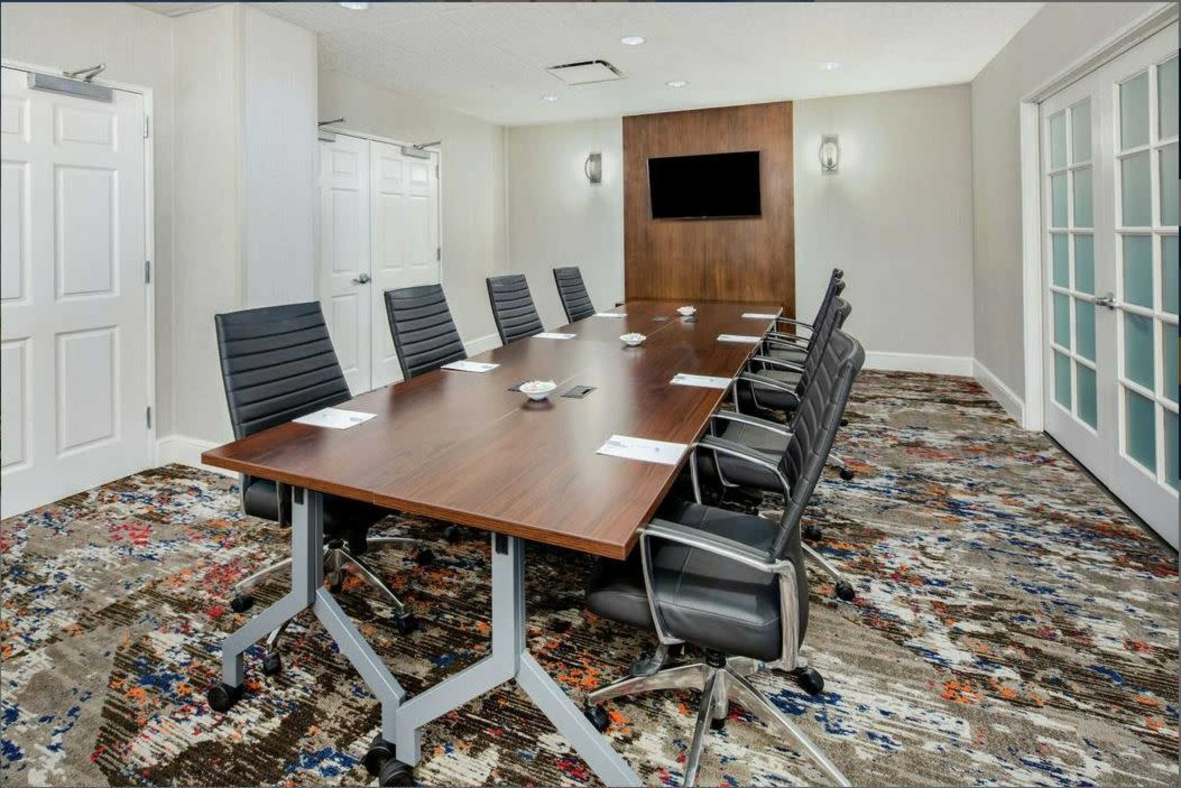 A modern conference room features a long wooden table surrounded by black office chairs, with a wall-mounted screen and double doors at either end.