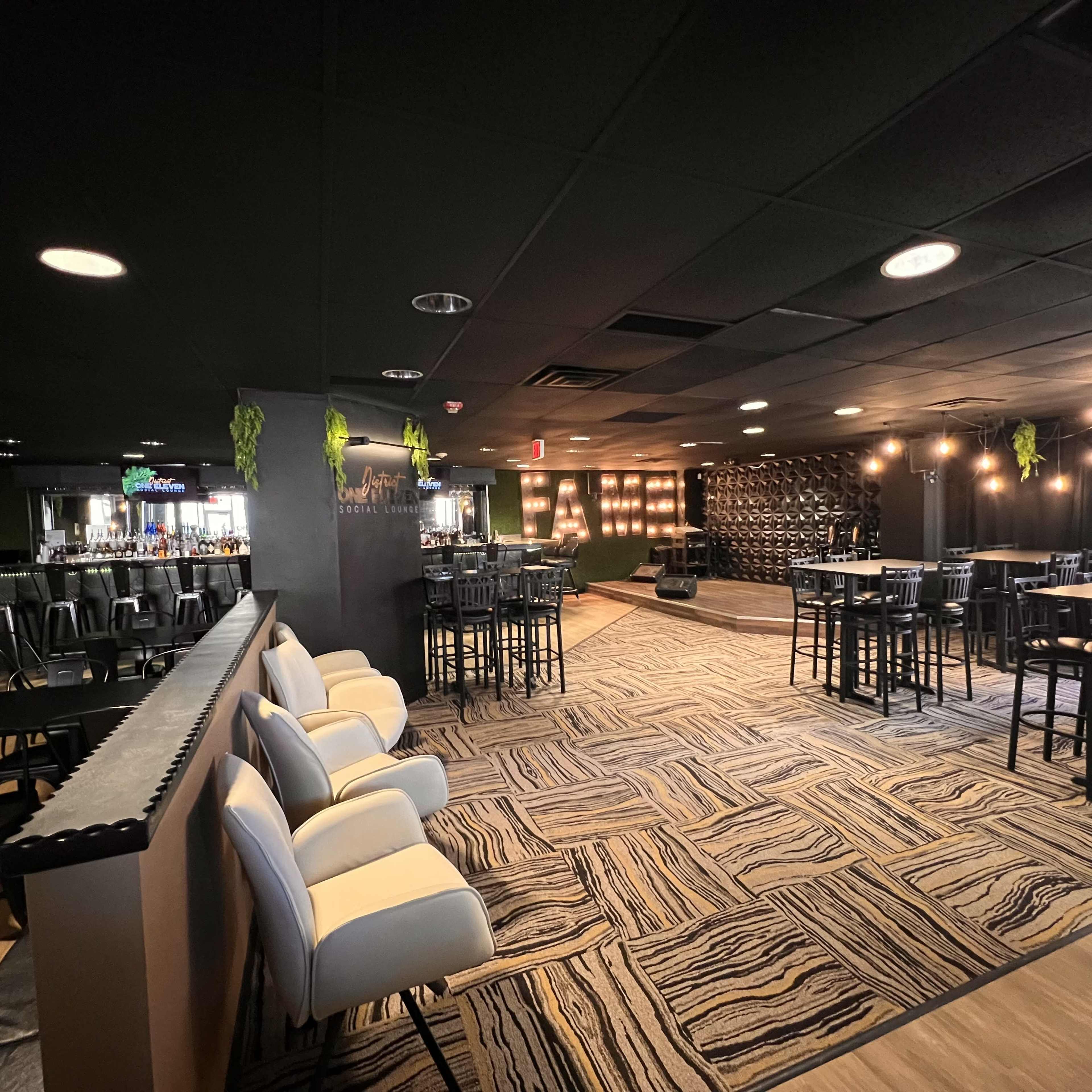 The image shows a modern lounge area with seating, bar tables, and a performance stage illuminated by large light-up letters.