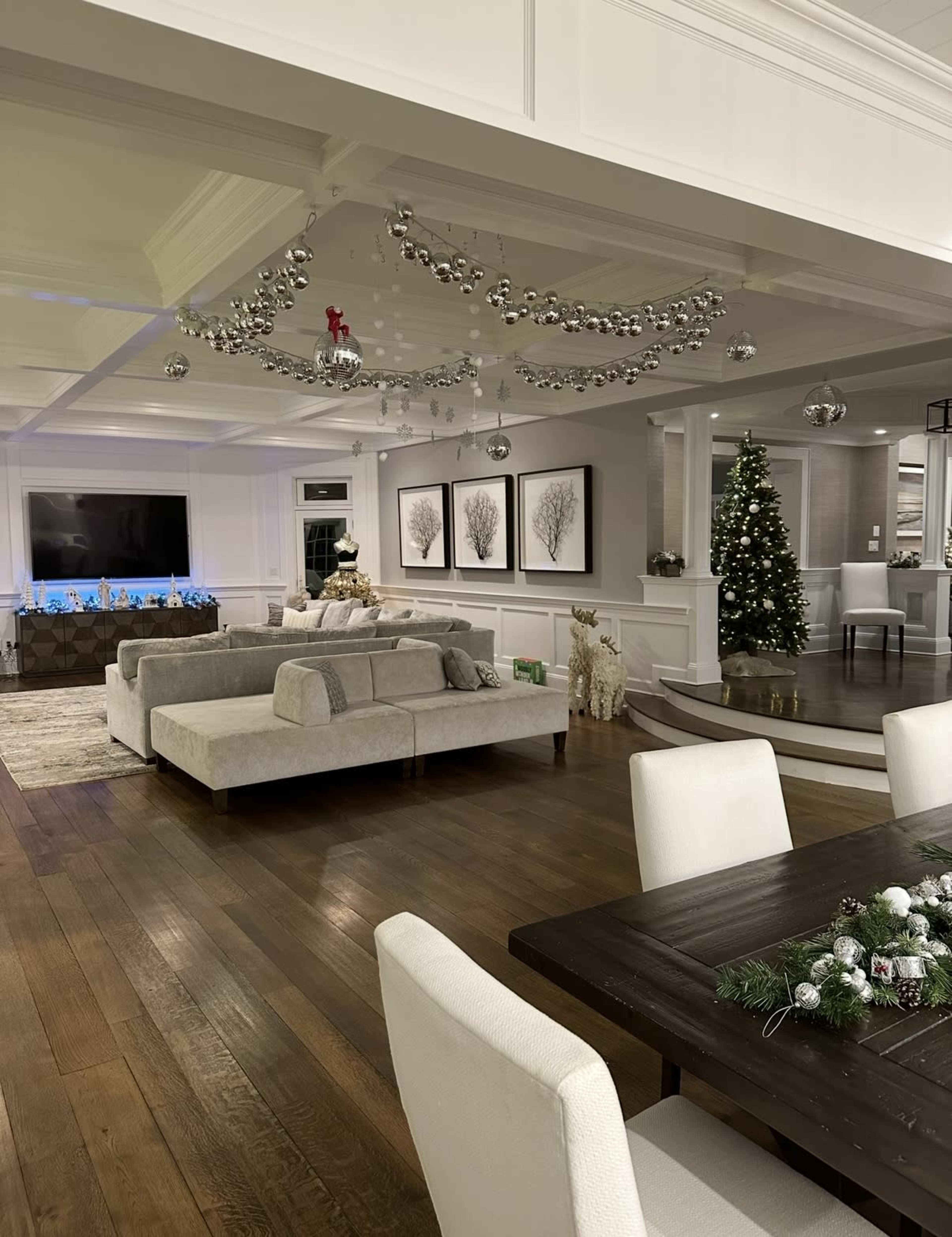 A spacious, elegantly decorated living area features a grey sectional sofa, a dining table with white chairs, and a Christmas tree, with silver decorations hanging from the ceiling.